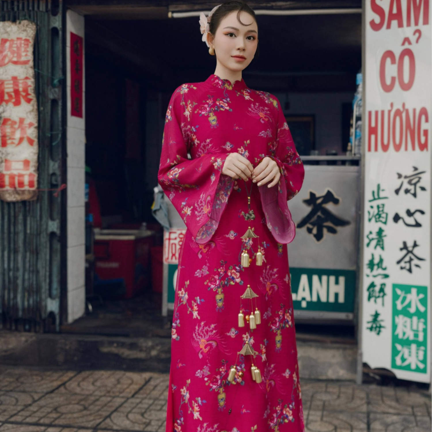 A woman standing in front of a store wearing a traditional Vietnamese magenta ao dai with gold and green floral patterns.