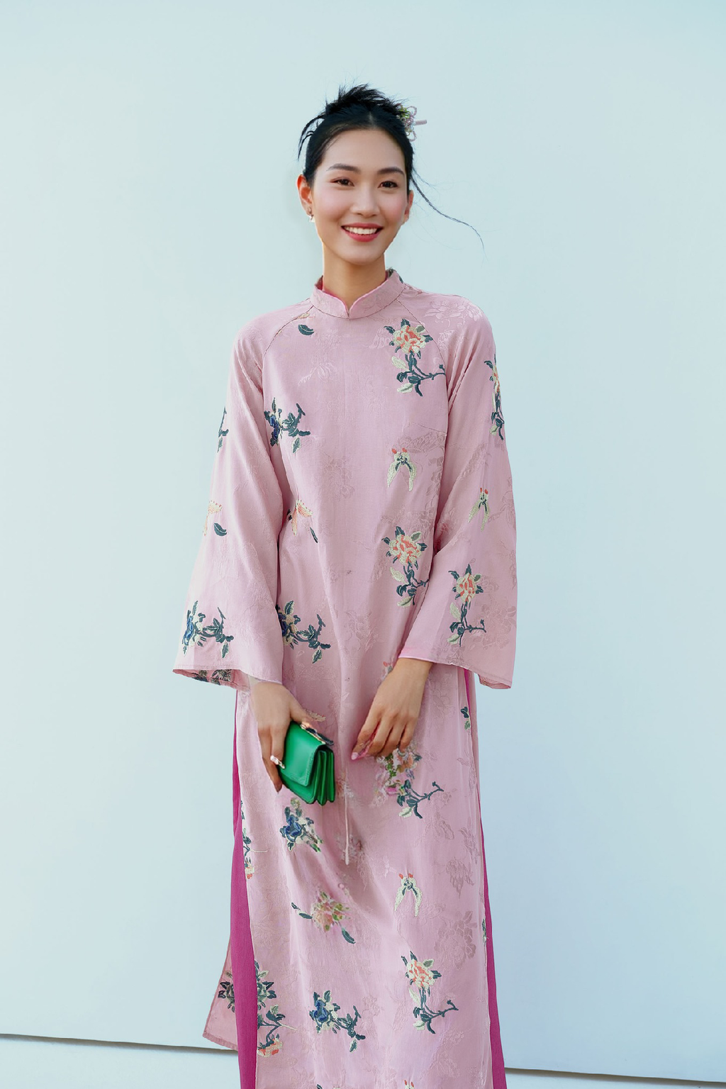 A person wearing a pink floral embroidered áo dài, holding a green purse, against a light background.