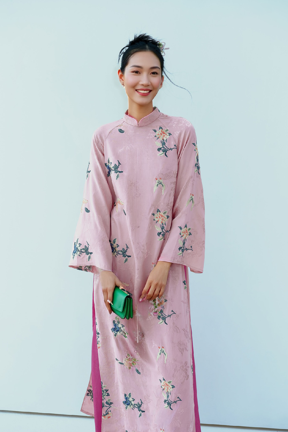 A person wearing a pink floral embroidered áo dài, holding a green purse, against a light background.