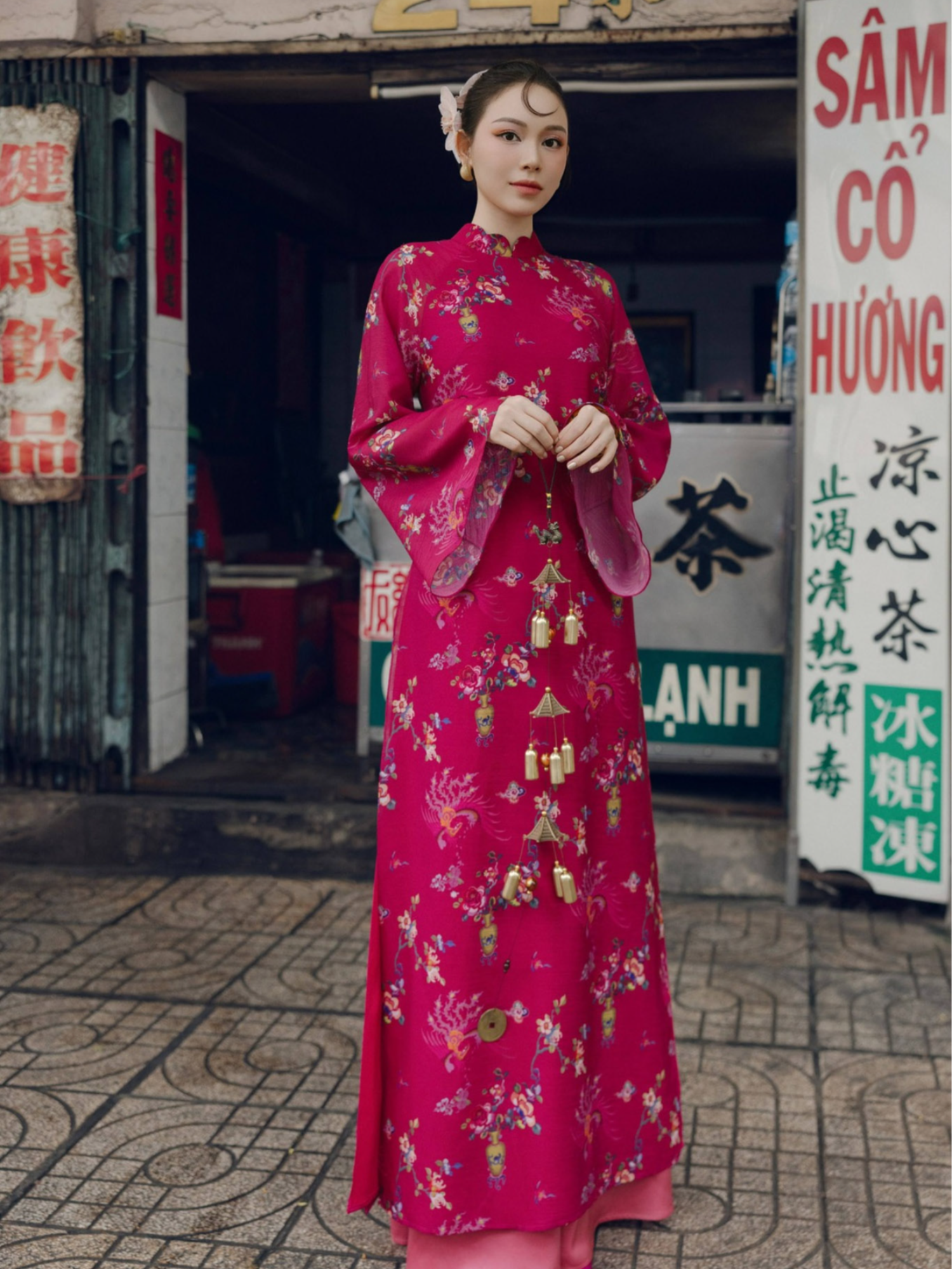 A woman standing in front of a store wearing a traditional Vietnamese magenta ao dai with gold and green floral patterns.