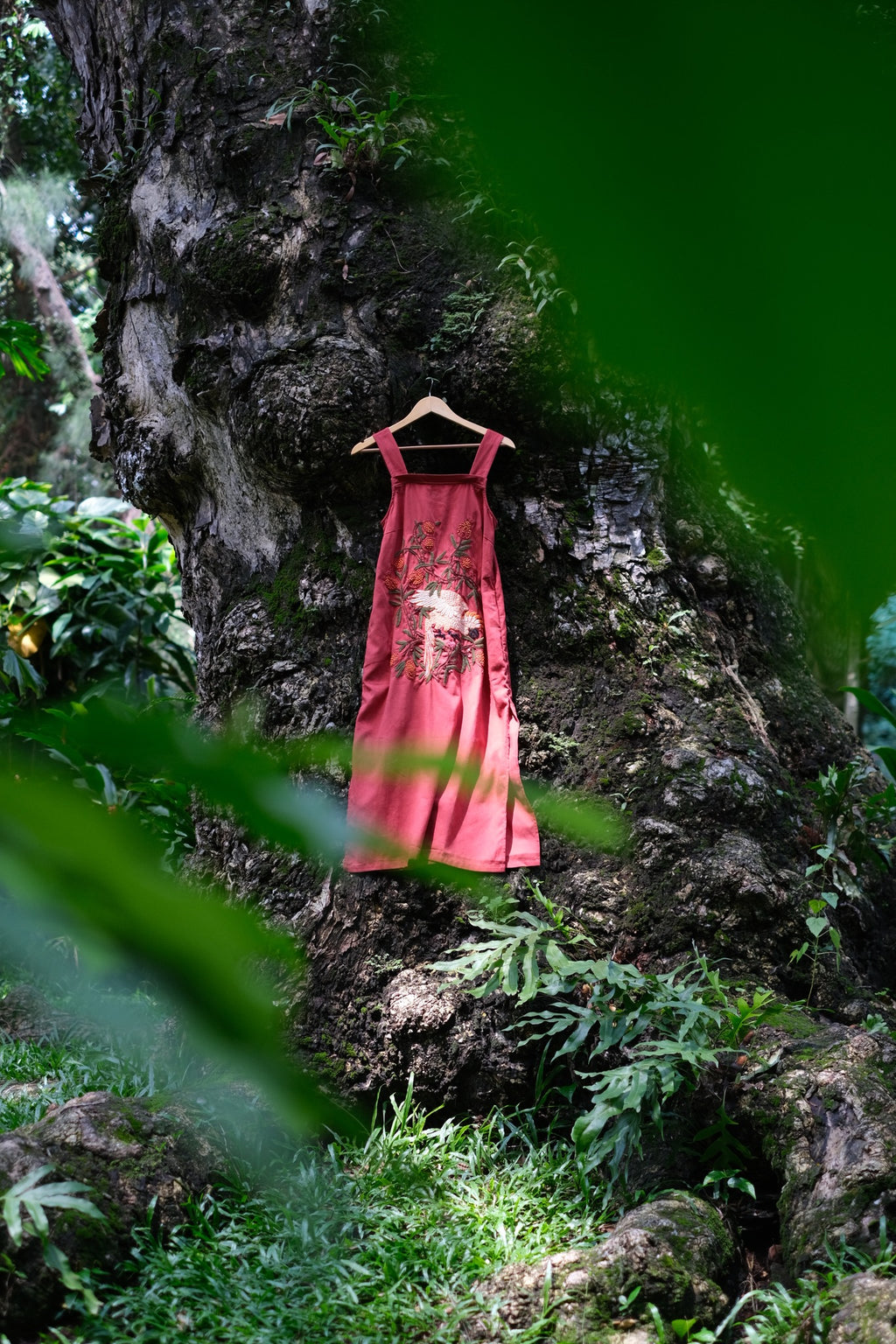 Scarlet Canopy hand-embroidered linen dress in terracotta red featuring white bird and lychee branch motifs, against a tree