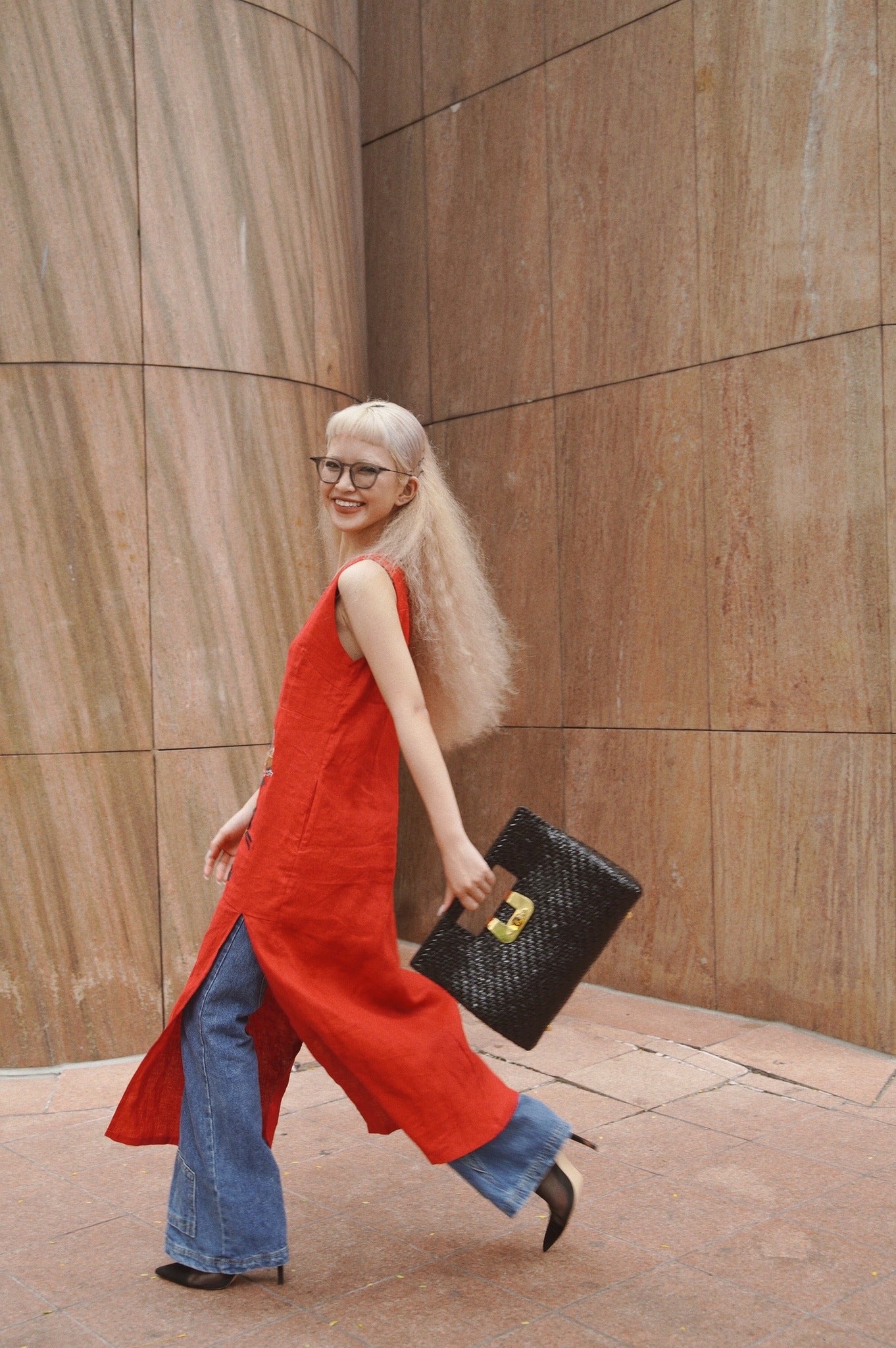 Woman in a sleeveless red tunic dress over flared blue jeans, carrying a black woven handbag and wearing black heels, smiling while walking past a stone wall.