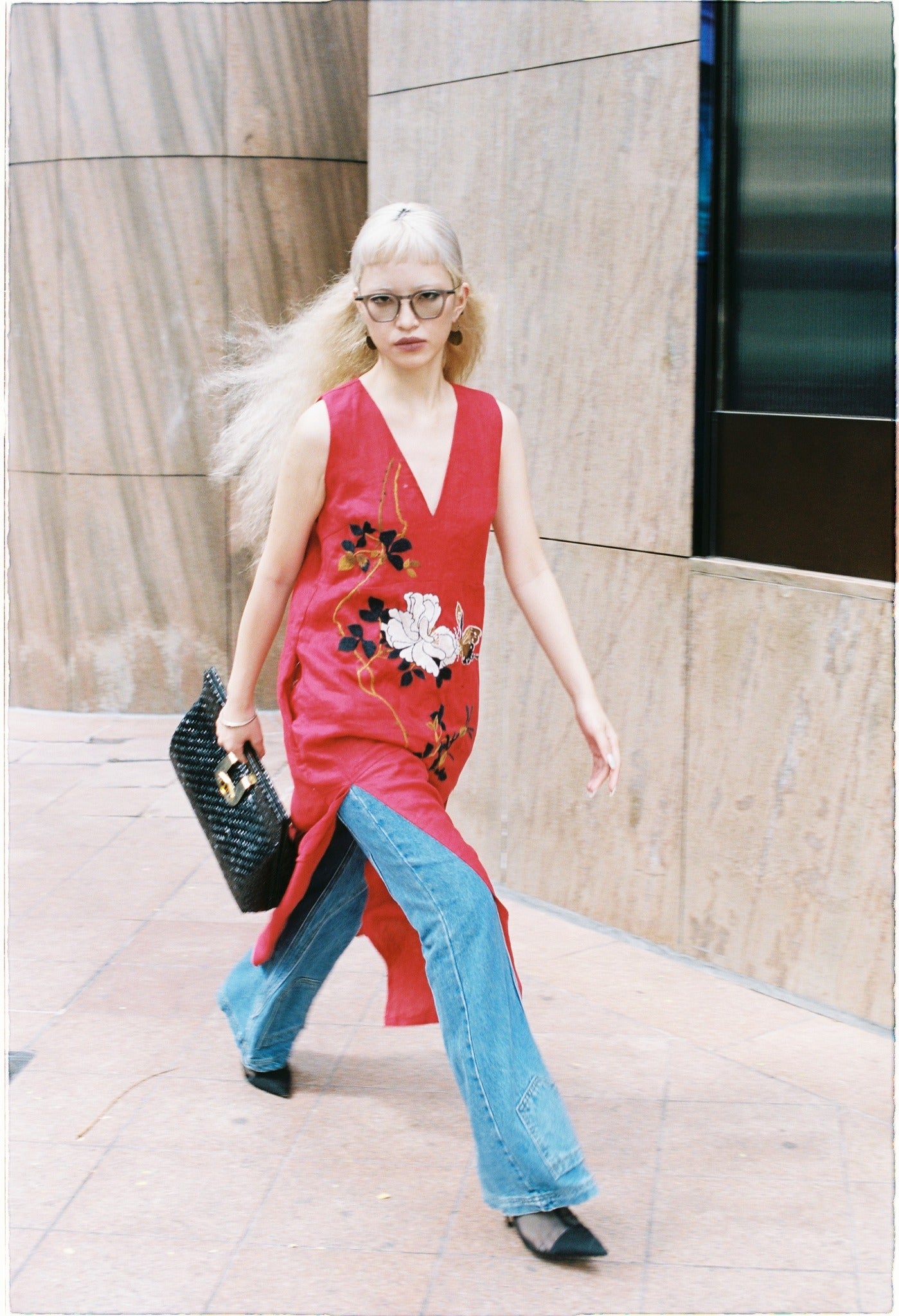 Woman walking confidently in a red sleeveless floral tunic layered over flared blue jeans, holding a black woven handbag.