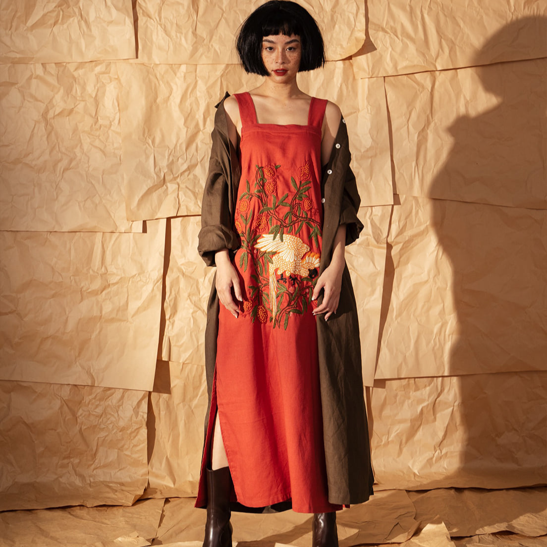 Scarlet Canopy hand-embroidered linen dress in terracotta red featuring white bird and lychee branch motifs, worn by woman against textured paper backdrop