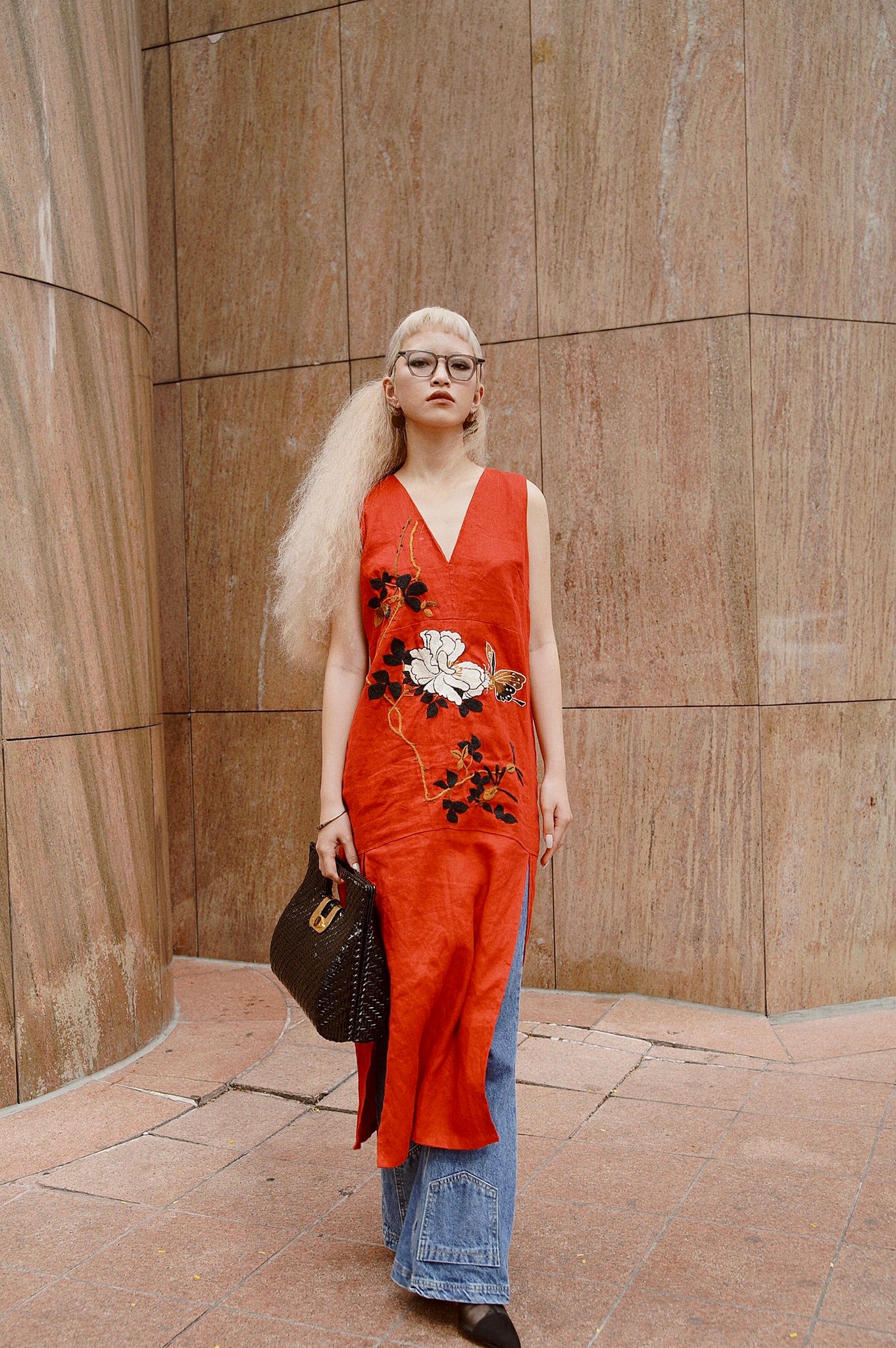 Woman wearing a red sleeveless dress with floral embroidery over wide-leg blue jeans, holding a textured black handbag.