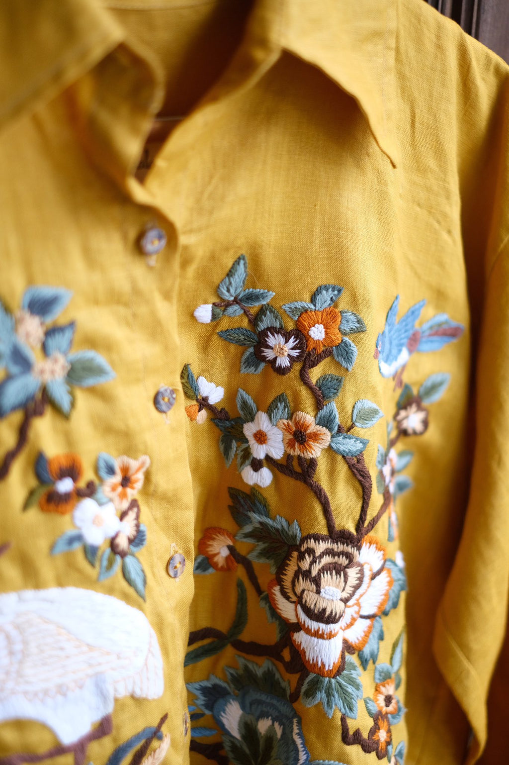 Close-up of a mustard yellow shirt featuring detailed floral embroidery in shades of blue, white, orange, and brown.