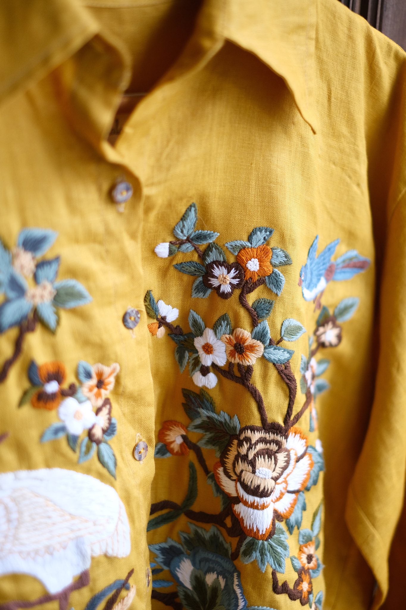 Close-up of a mustard yellow shirt featuring detailed floral embroidery in shades of blue, white, orange, and brown.