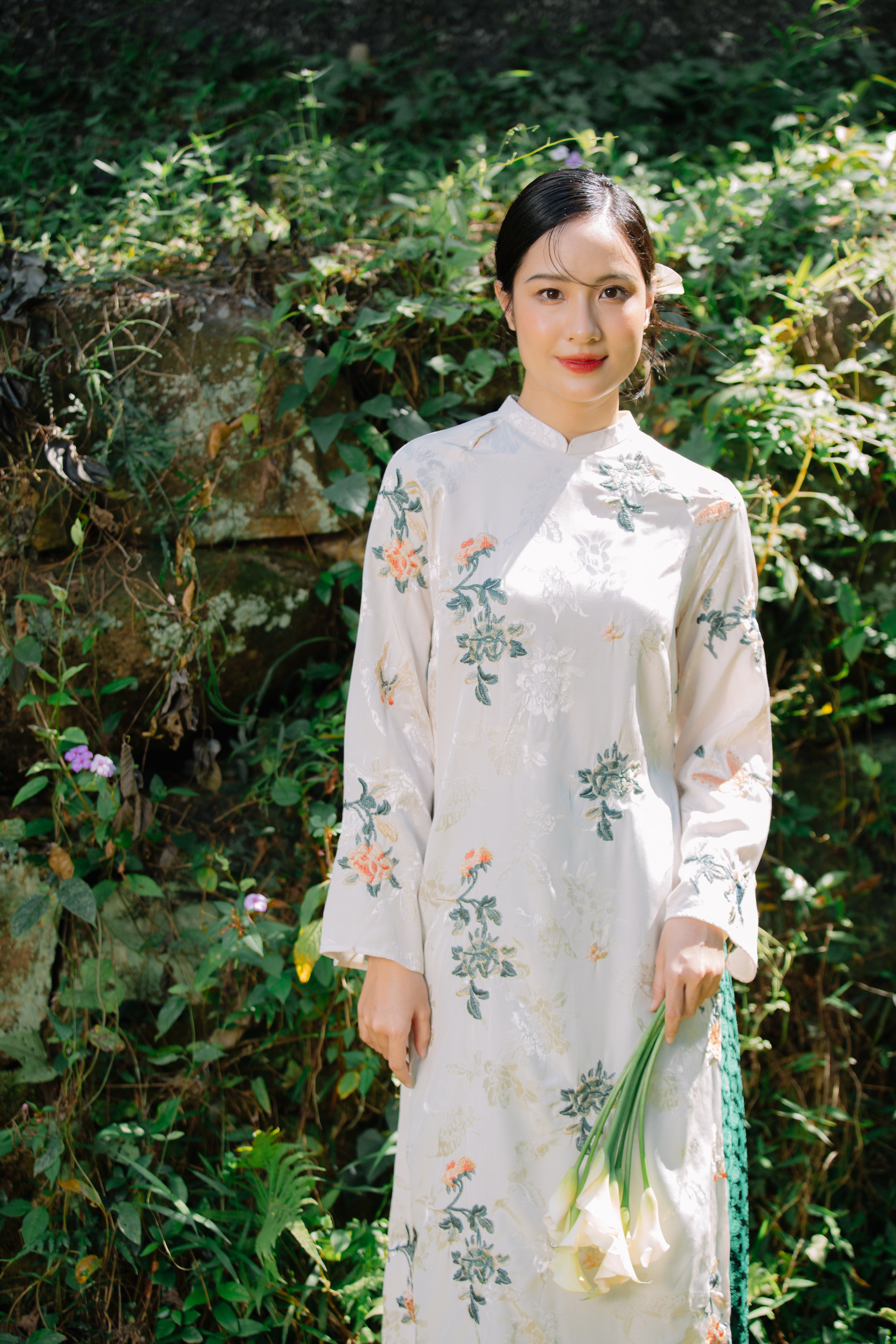 A person wearing a floral embroidered white dress, holding a small bouquet of white flowers, stands in front of a leafy green background.