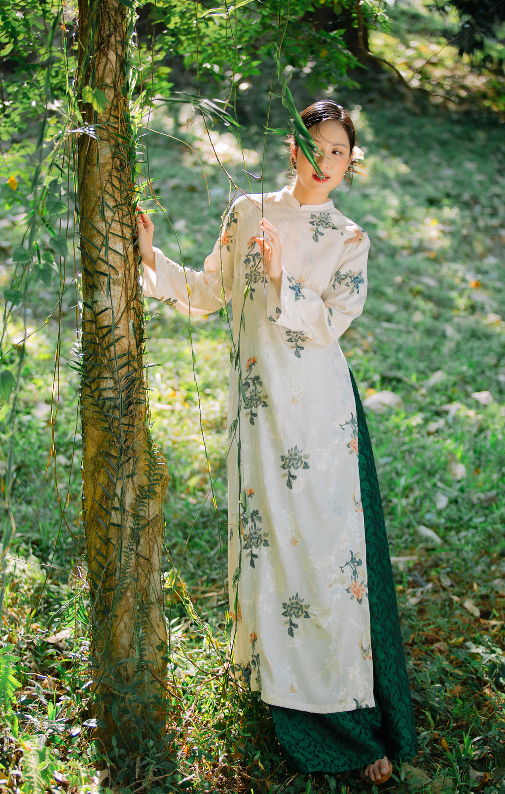 Woman in a floral-patterned dress stands beside a tree in a sunlit forest, surrounded by greenery.