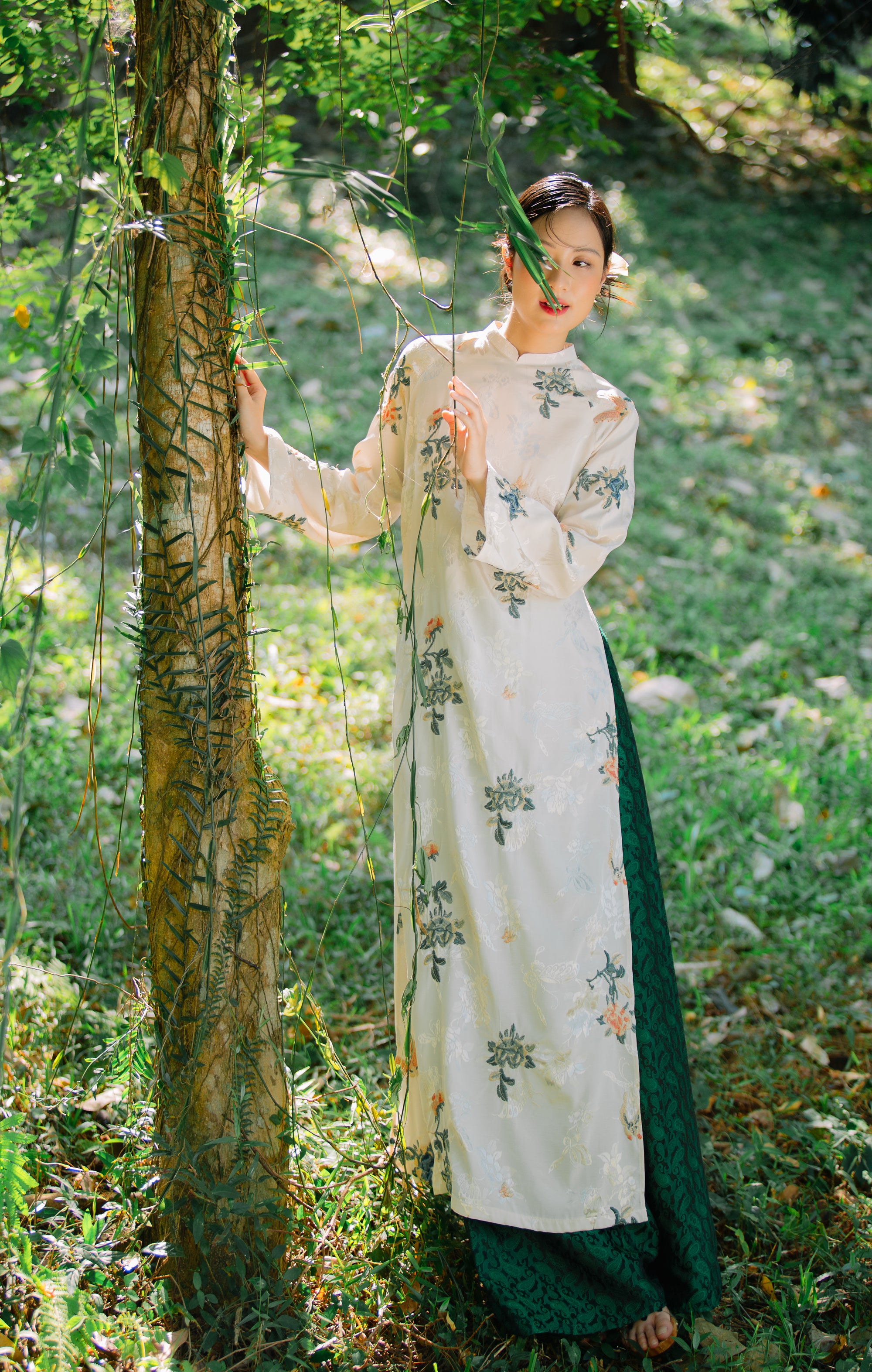 Woman in a floral-patterned dress stands beside a tree in a sunlit forest, surrounded by greenery.