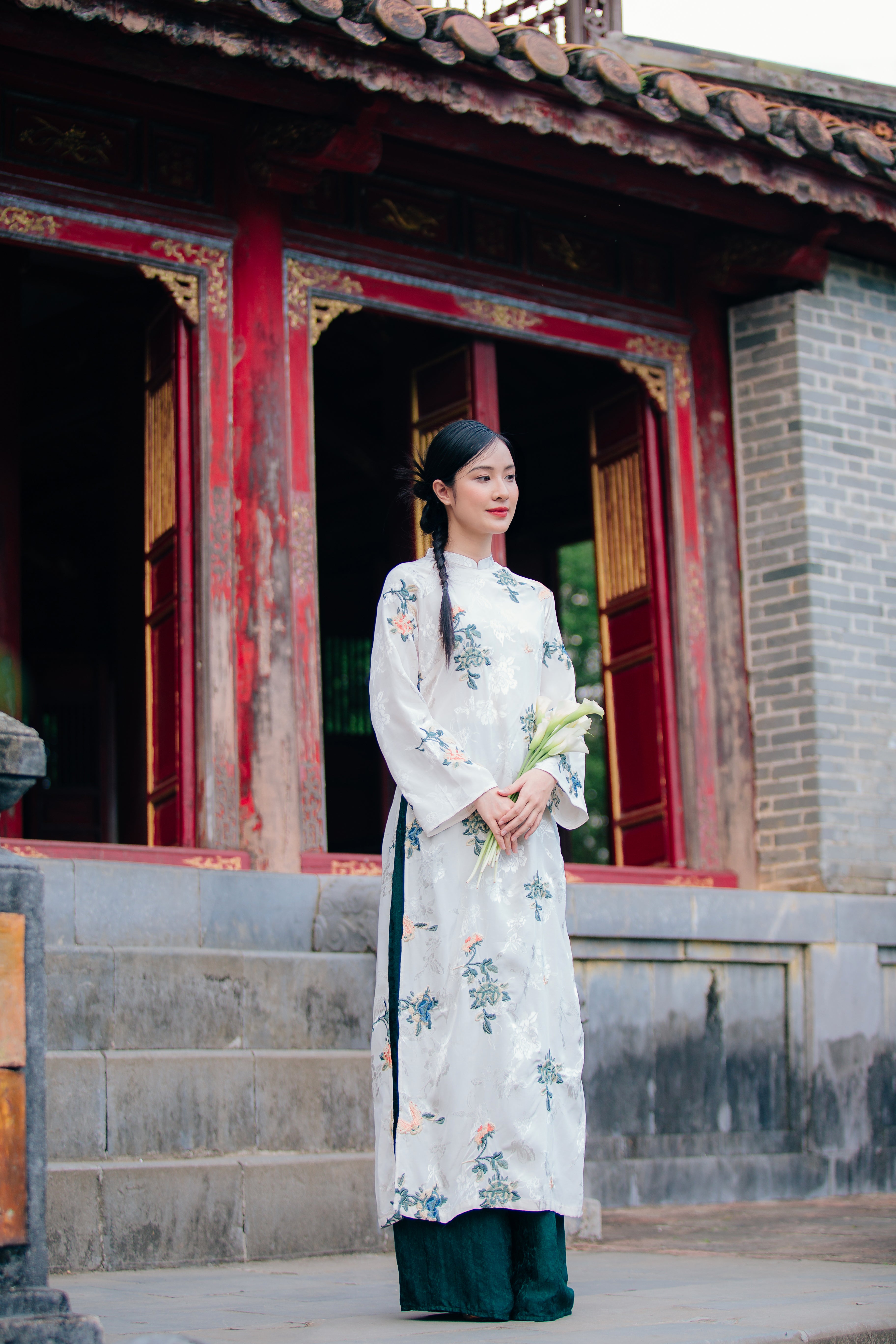 A woman stands gracefully on stone steps in front of a traditional building wearing a long floral dress and holding white flowers.
