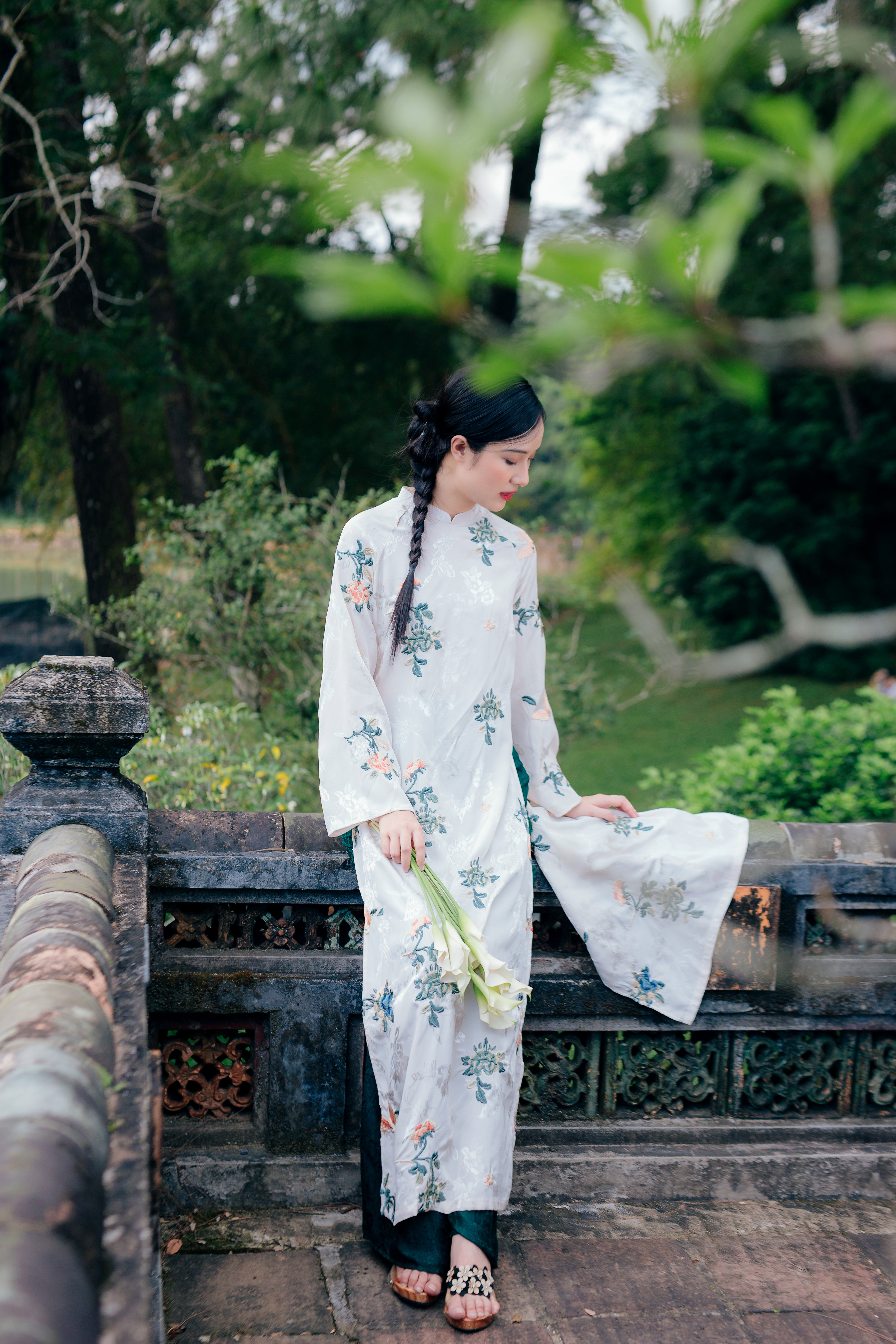 A woman in a white floral traditional dress stands by an ornate stone railing in a lush garden, holding white flowers.