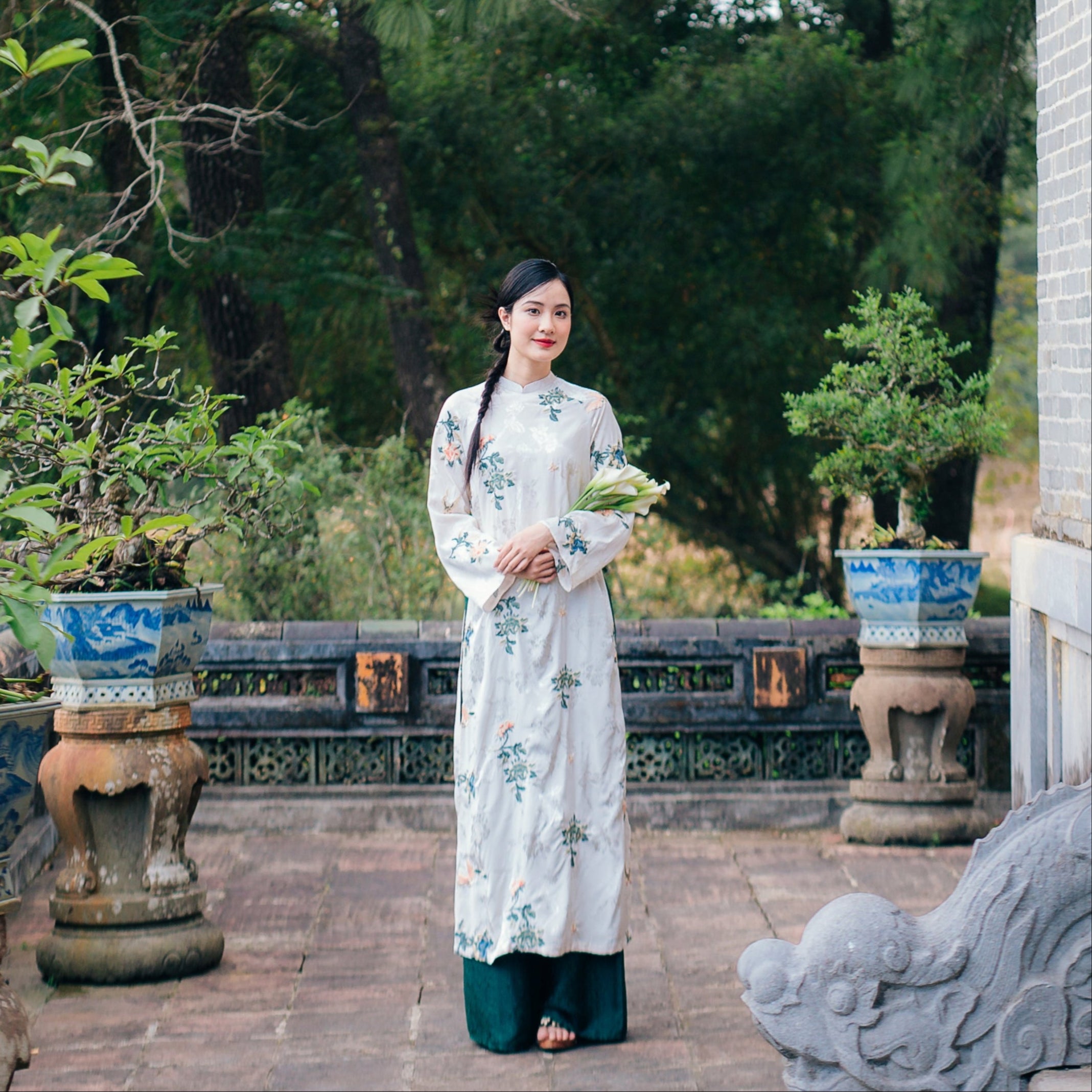 A woman in a floral embroidered traditional dress stands outdoors near potted plants, holding white flowers.