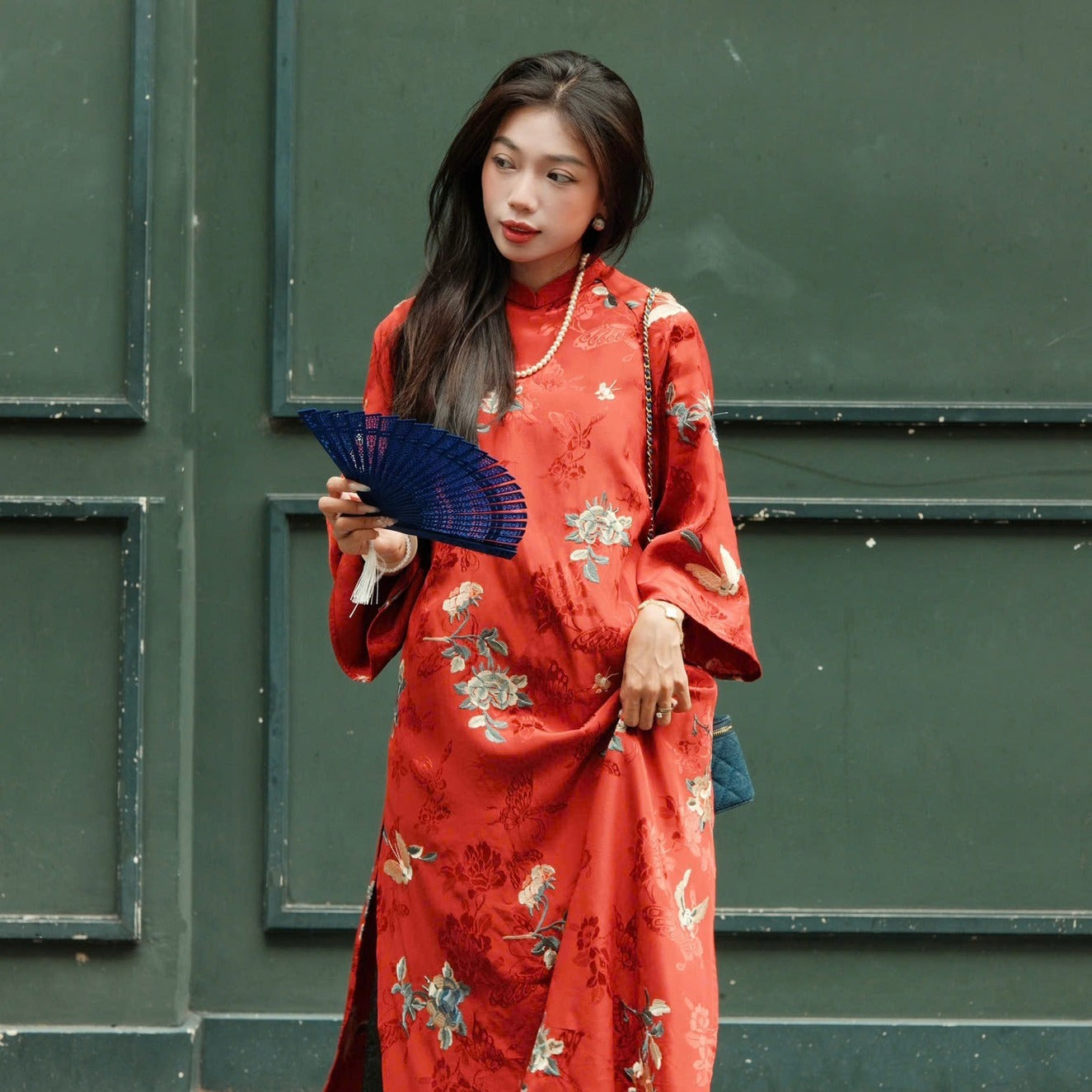 A person wearing a red floral dress with traditional patterns, holding a blue fan.