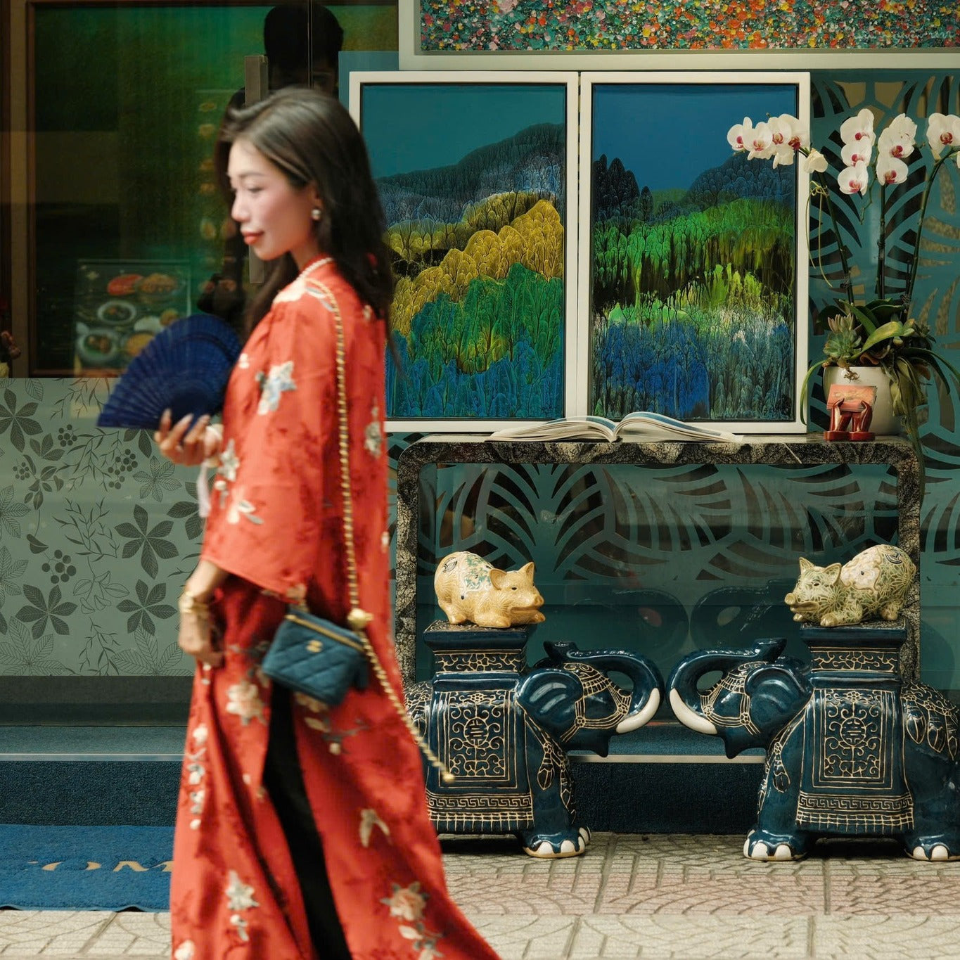A woman in a red floral kimono walks past a decorative display featuring golden animal figurines, elephant statues, and vibrant landscape paintings.
