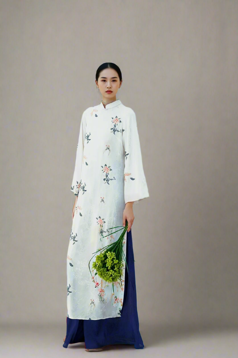 A person wearing a long, white, floral-embroidered traditional dress with blue pants, holding a bouquet of green flowers.