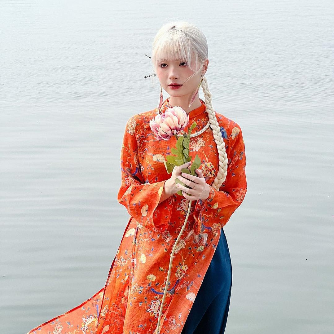 A person wearing a traditional orange embroidered outfit holds a pink lotus flower by a calm body of water.