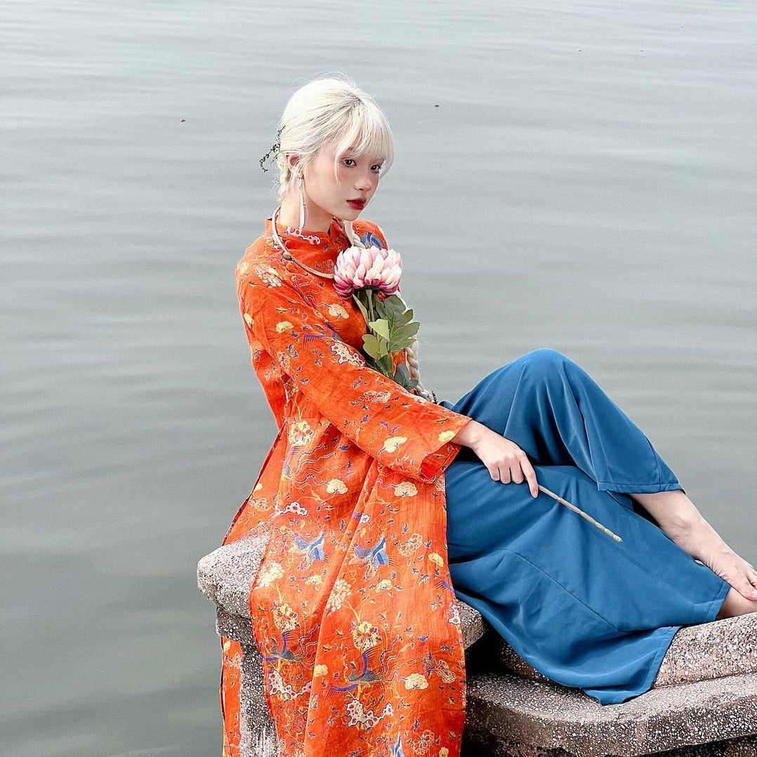 A person in a vibrant orange traditional outfit sits near a body of water while holding a flower, with relaxed posture and serene surroundings.