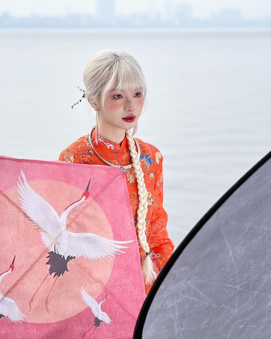 A person in an orange floral outfit holds a pink object with crane illustrations, standing by a body of water.