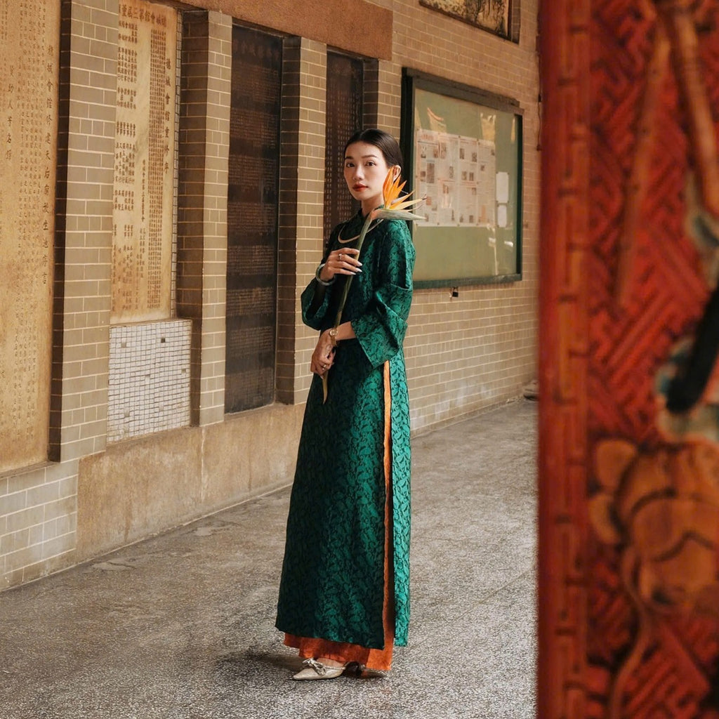A person in a long, green traditional dress holds a bird of paradise flower, standing in a corridor lined with framed displays and beige tiles.