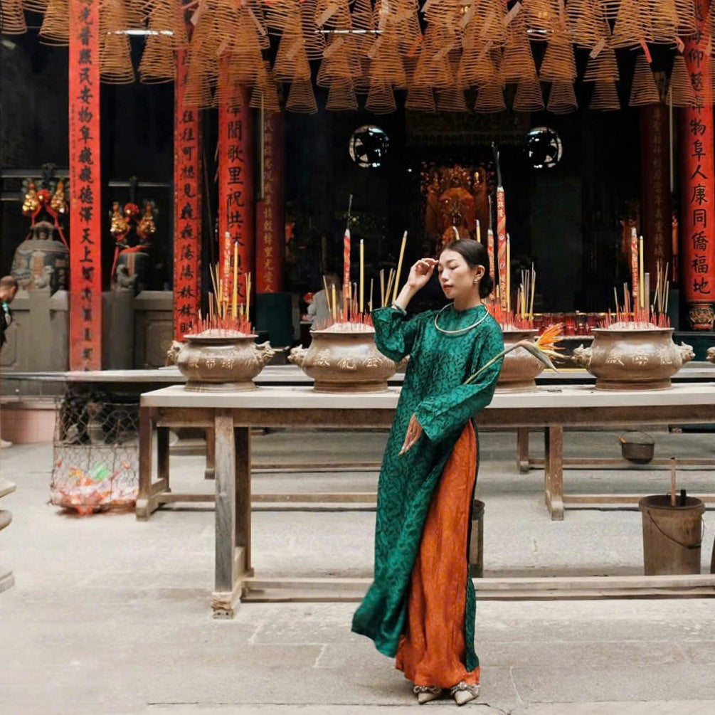 A woman wearing a traditional green and orange dress stands in an ornate temple with incense pots and hanging coils in the background.