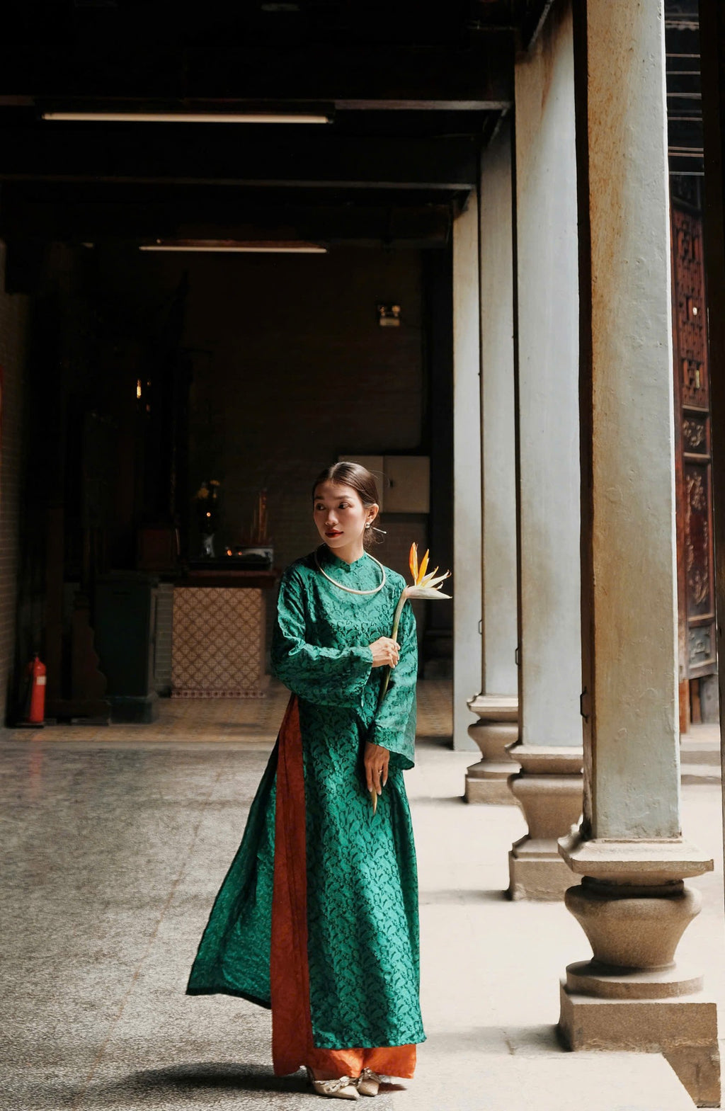 A person in a green patterned traditional dress, holding a tropical flower, stands in an ornate indoor space with tiled floors and large columns.