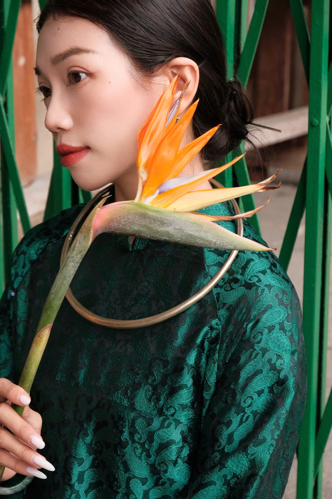 A woman in a green patterned dress holds a vibrant orange bird of paradise flower.