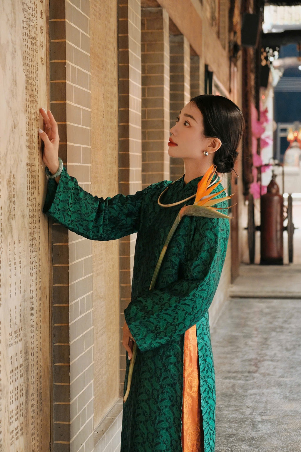 A person in an elegant green traditional dress stands in a hallway, gently touching a wall with text, holding a vibrant orange bird of paradise flower.