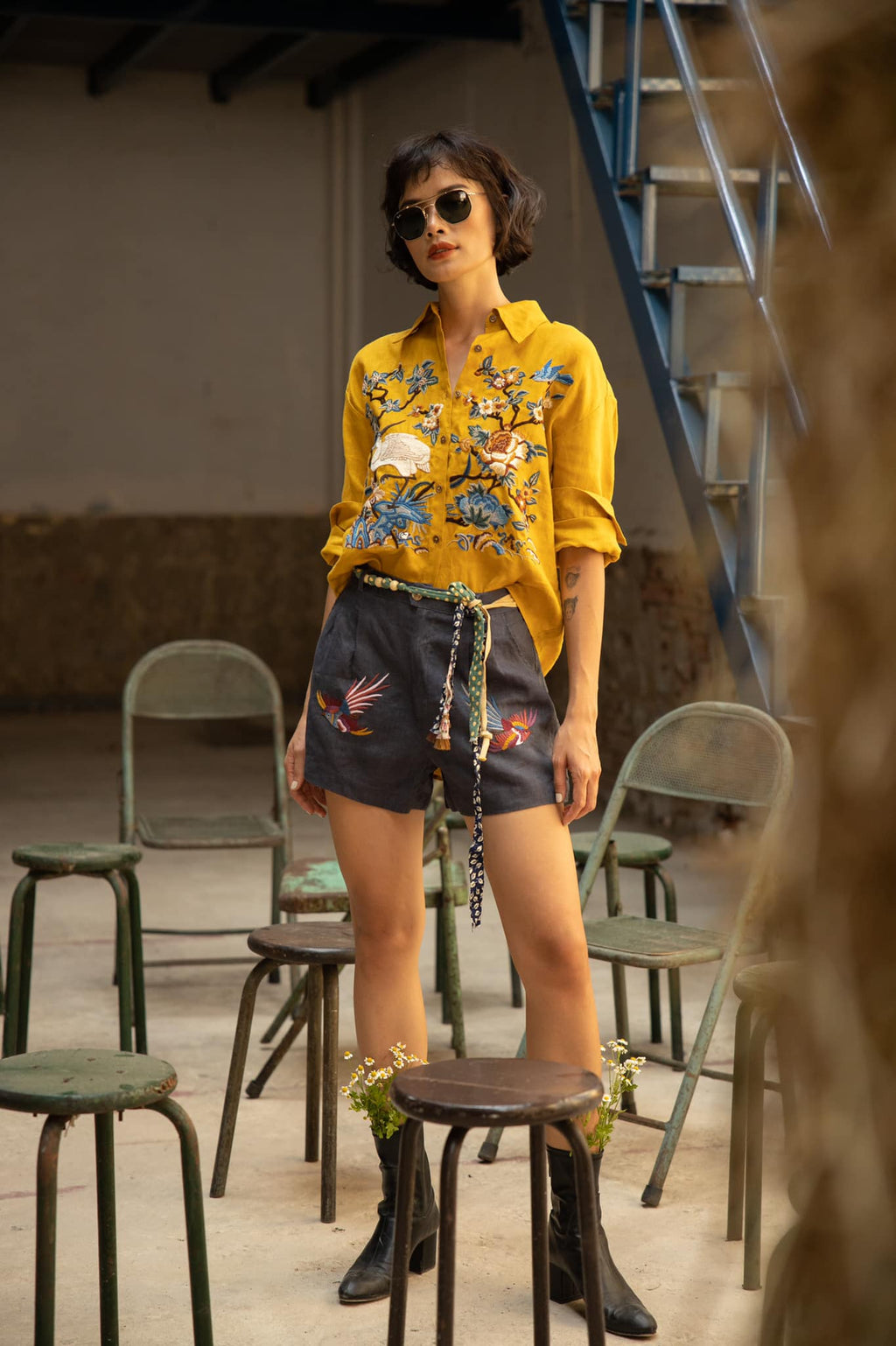 Woman wearing a mustard yellow embroidered shirt with floral and bird motifs, paired with dark shorts featuring bird designs, a beaded belt, black boots, and sunglasses, standing among vintage stools in an industrial setting.