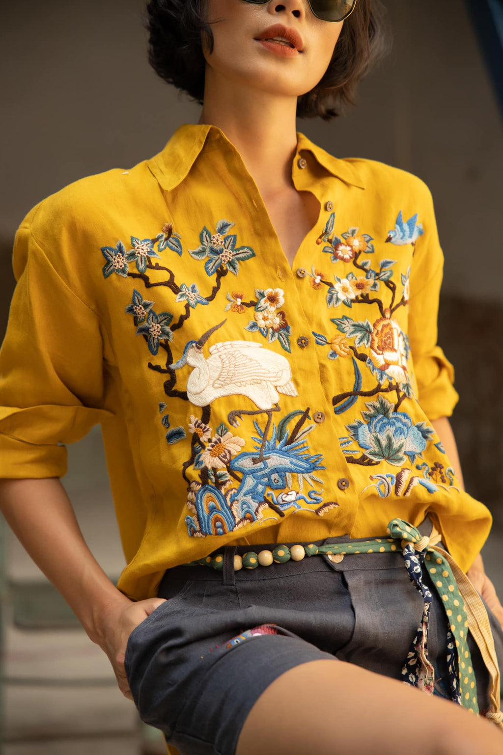 Woman wearing a vibrant mustard yellow blouse with intricate floral and bird embroidery, paired with dark high-waisted shorts and a decorative belt.