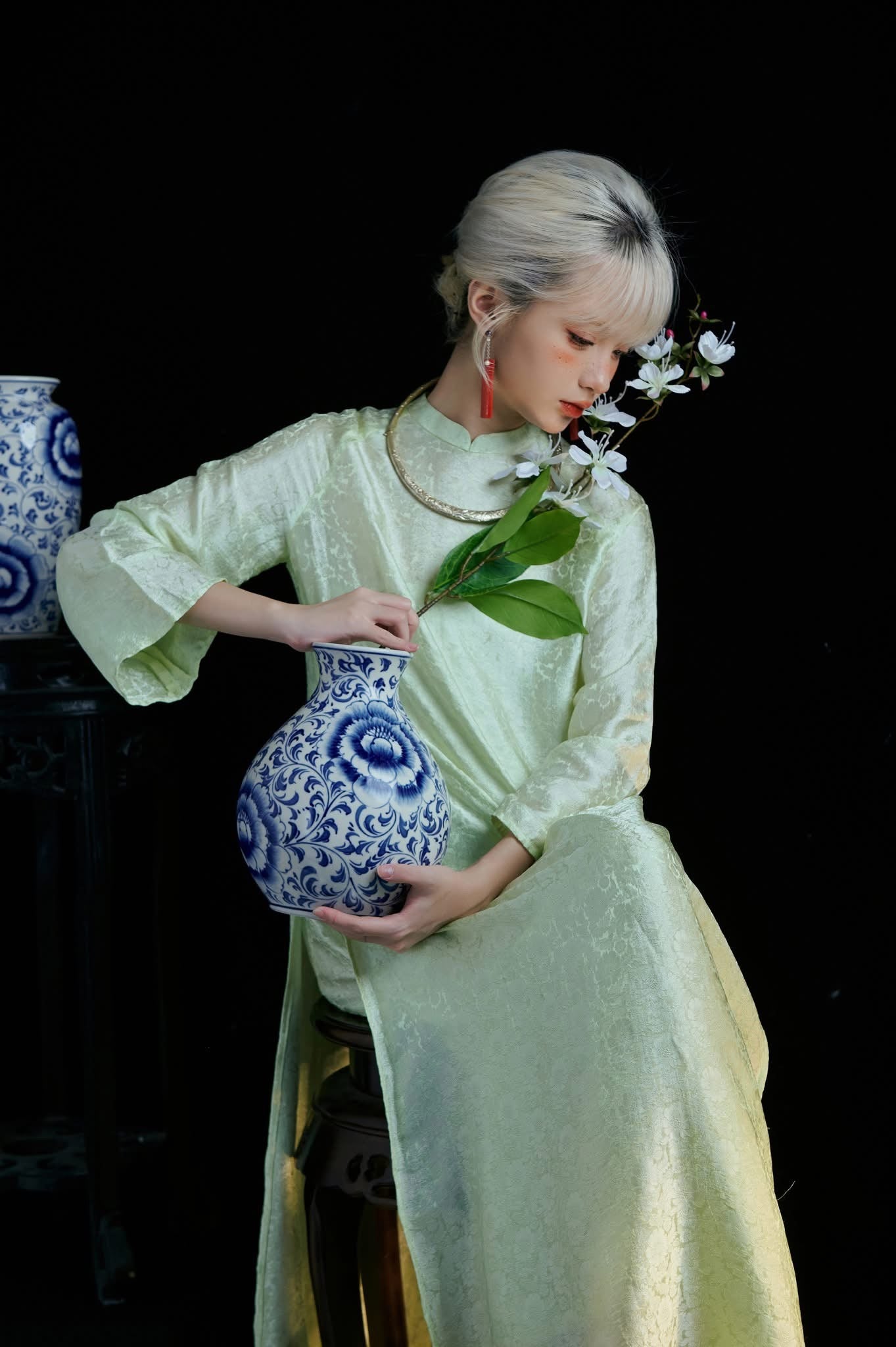 A person in a light green dress holding a blue and white floral vase with flowers, set against a dark background.