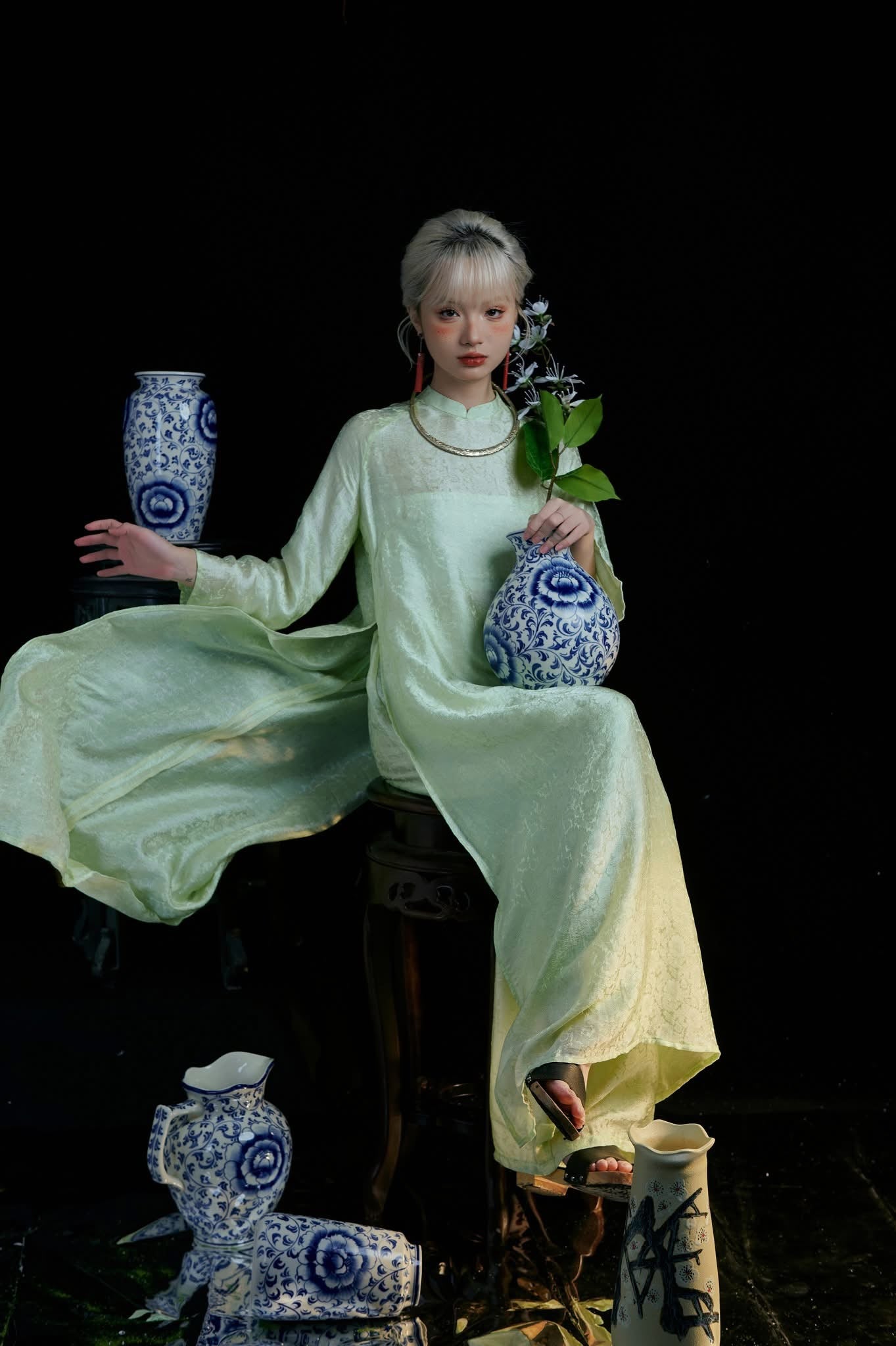 A person in a flowing light green dress sits among decorative blue and white vases, holding one with a flower. The background is dark, highlighting the porcelain patterns and delicate attire.
