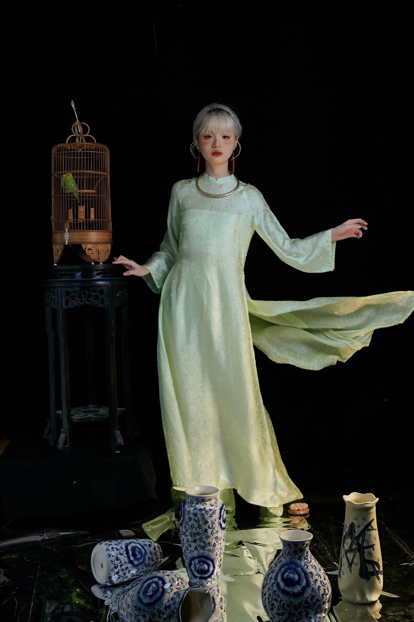 A person in a flowing, light green dress stands beside a birdcage on a dark wooden stand. Several blue and white porcelain vases are placed on the ground around them. The background is black, highlighting the scene.