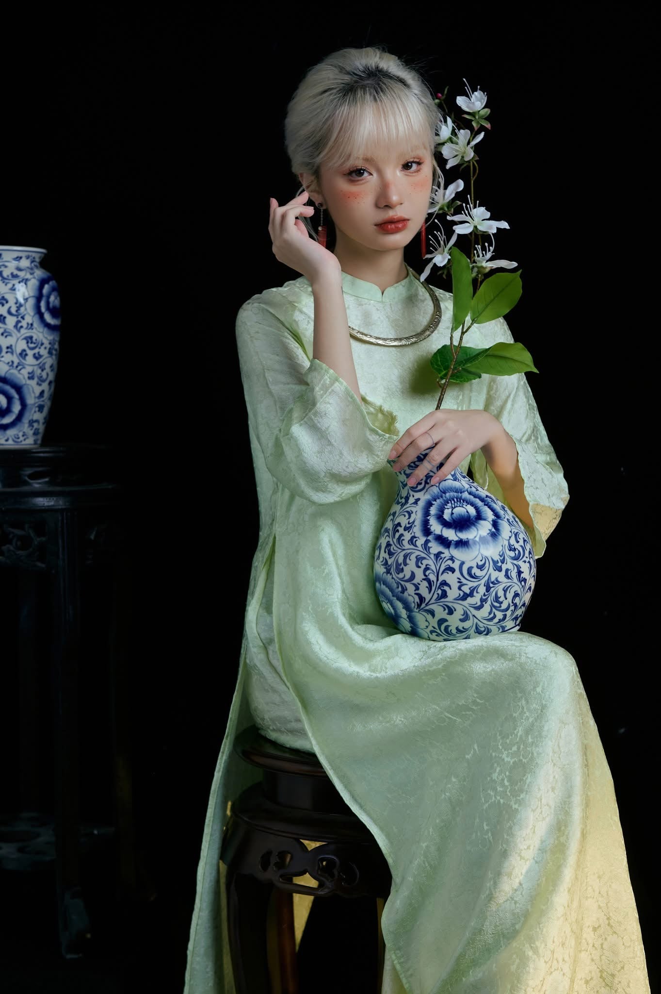 A person in a light green dress holds a blue and white floral-patterned vase with a branch of white flowers, sitting against a dark background. Another similar vase is partially visible on a stand.