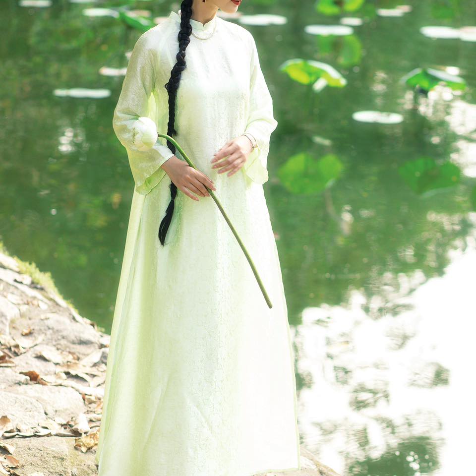 A woman in a long, light, flowing dress stands by a serene pond, holding a single white lotus.