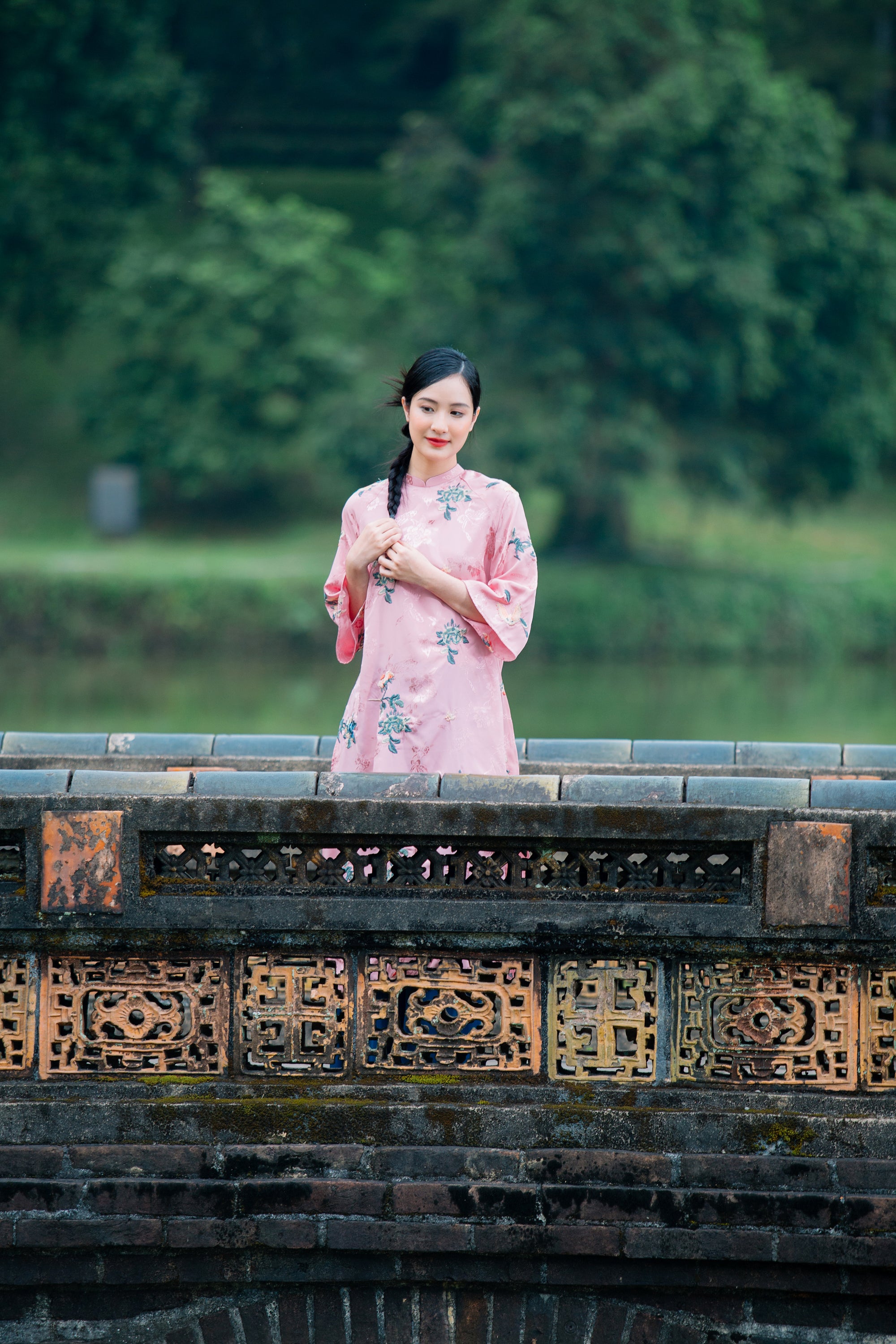 A person in a pink floral dress stands near a decorative stone railing, with blurred greenery in the background.