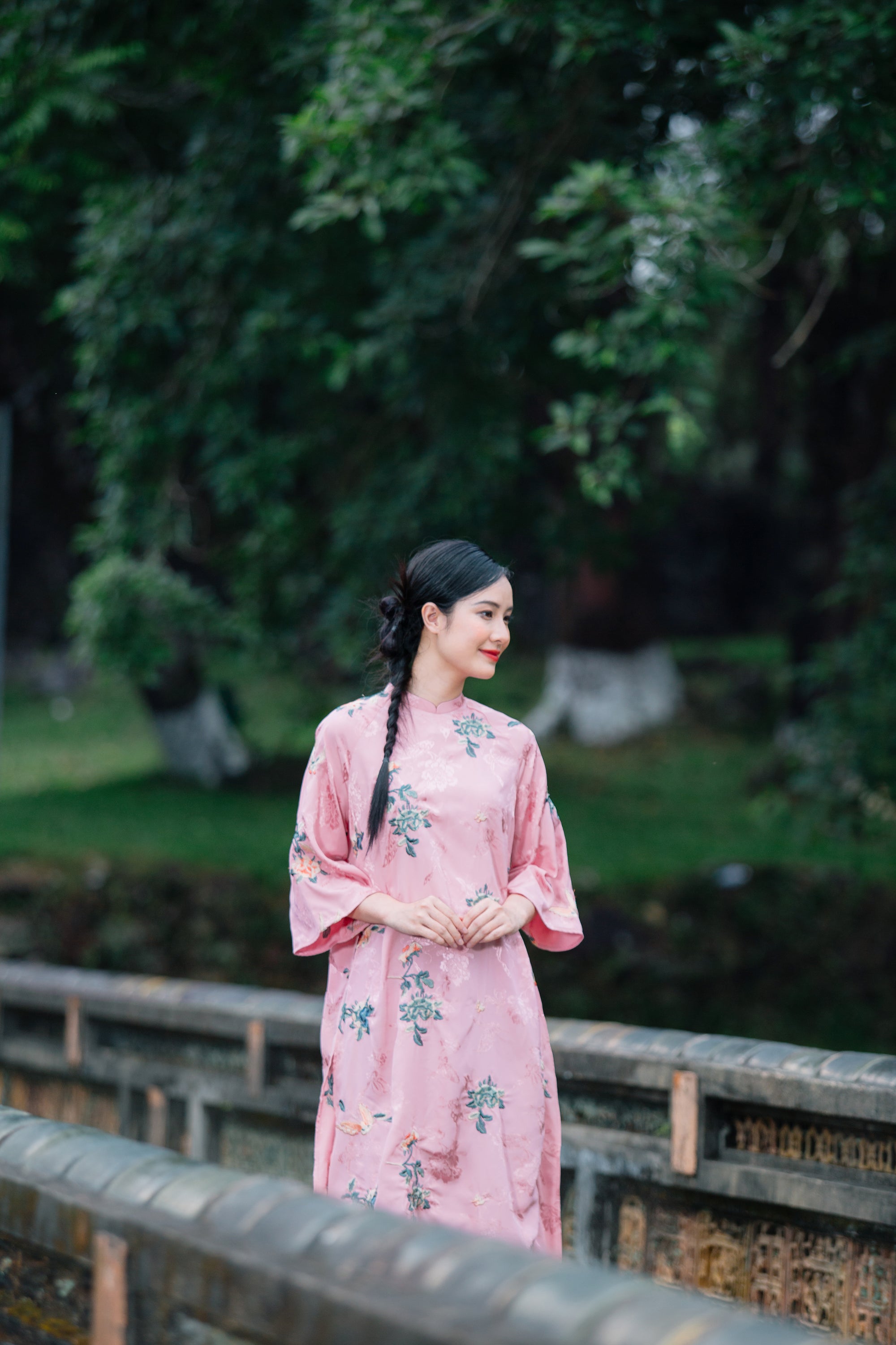 A woman in a pink floral dress standing outdoors on a wooden bridge, with green foliage in the background.
