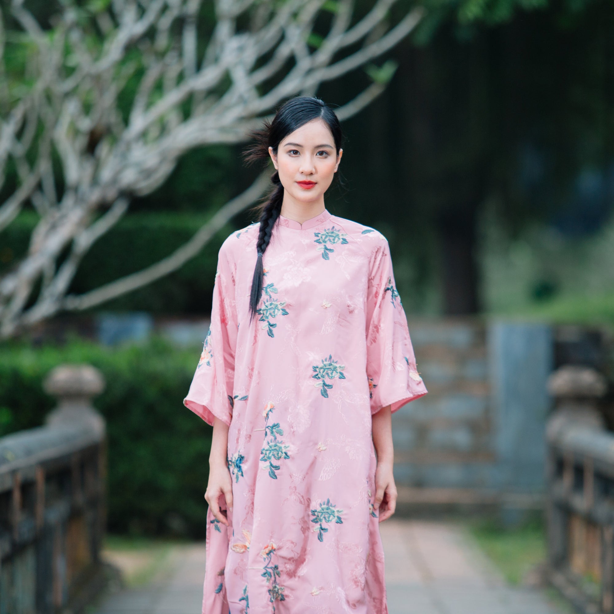 A person wearing a pink traditional dress with floral patterns, standing on a stone walkway with greenery in the background.