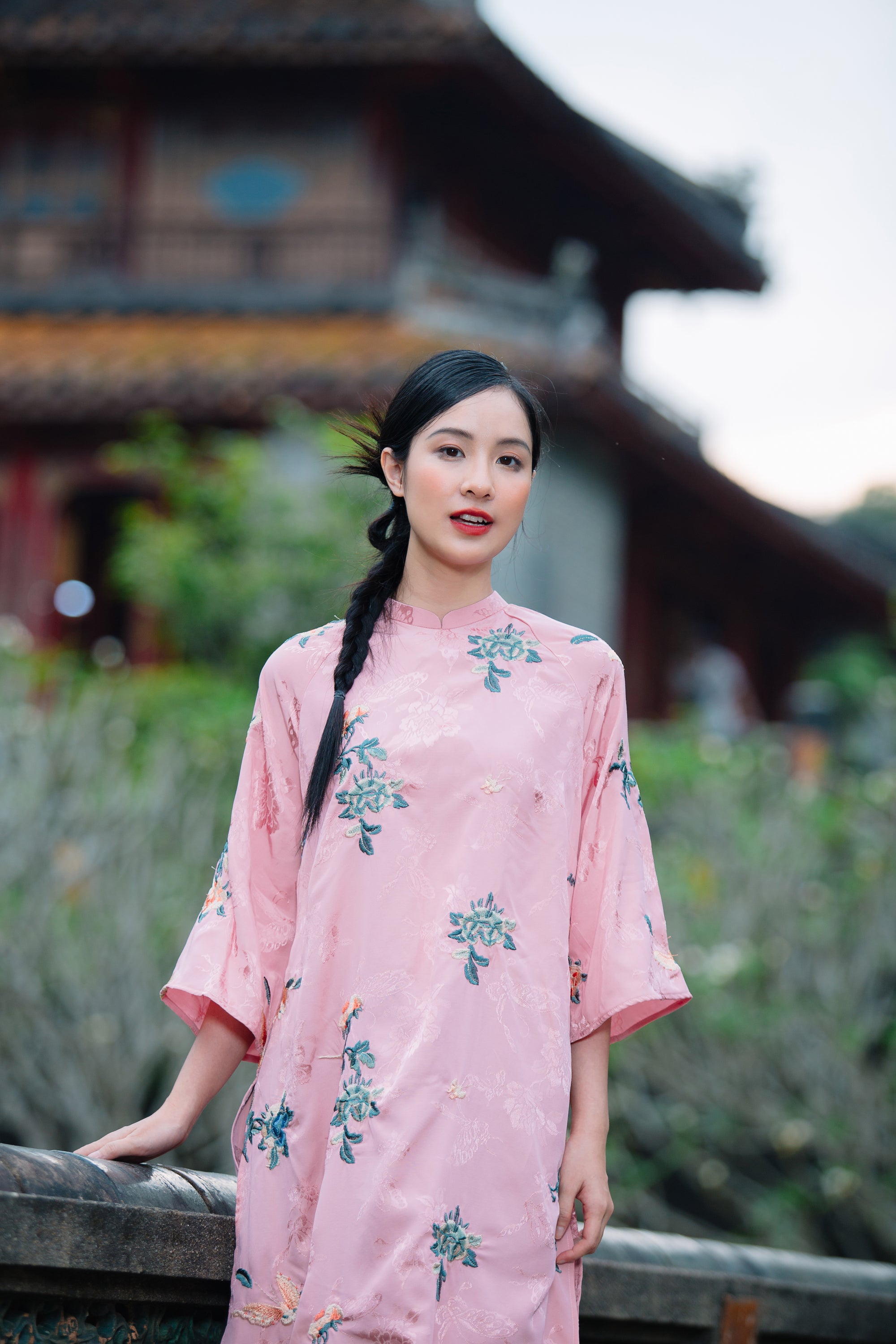 A person wearing a pink floral embroidered dress stands outdoors with a traditional building in the background.