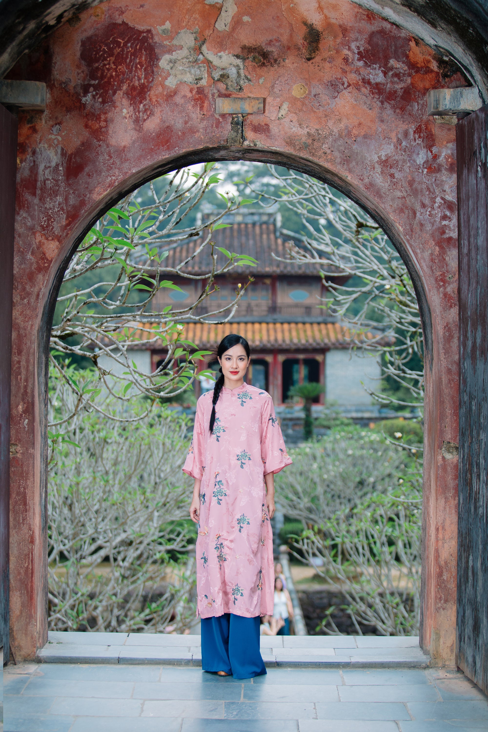 A person stands in a historic archway, wearing a pink floral dress and blue pants, with a serene garden and traditional building in the background.