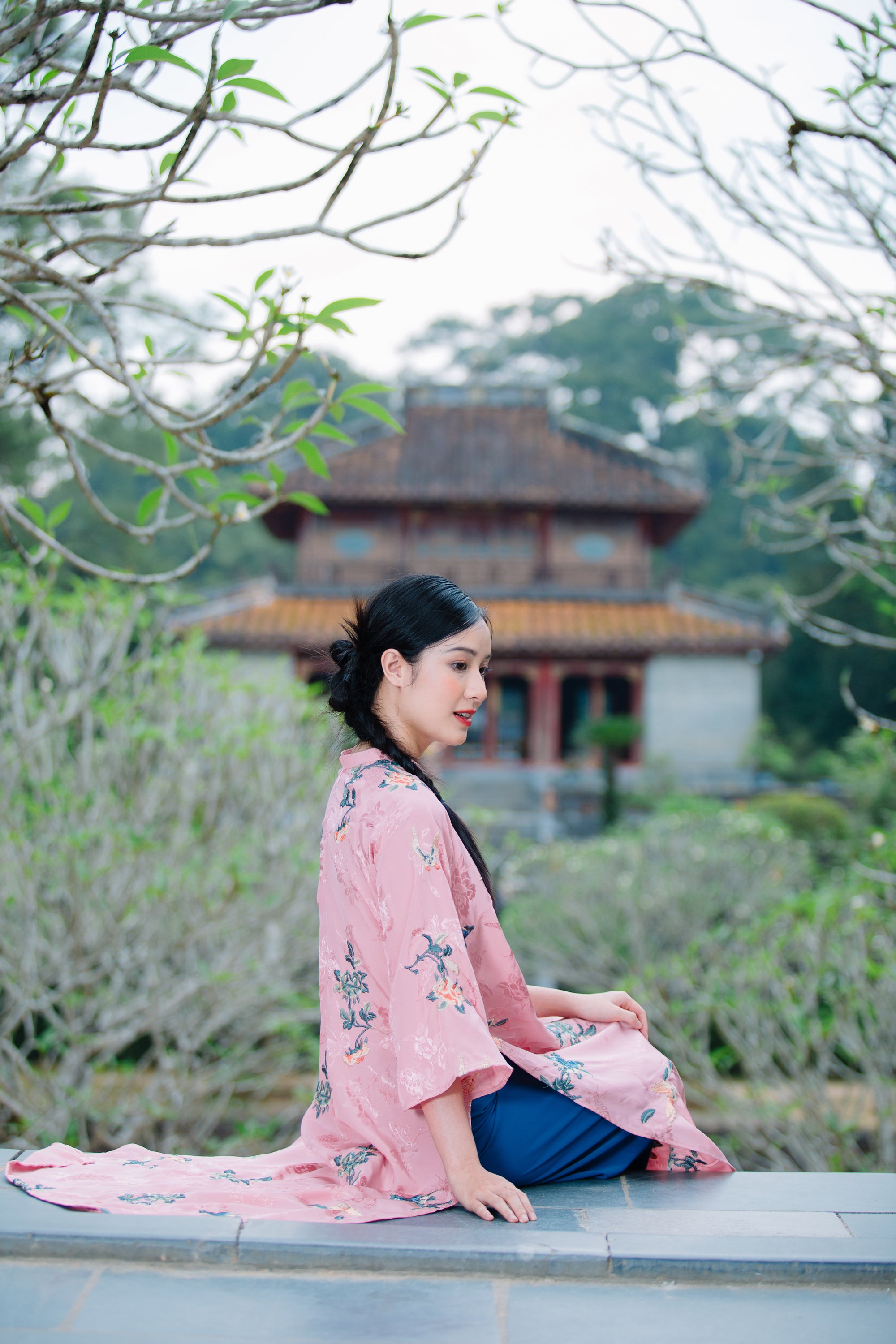 A person in a pink floral kimono sits gracefully with a traditional building and lush greenery in the background.