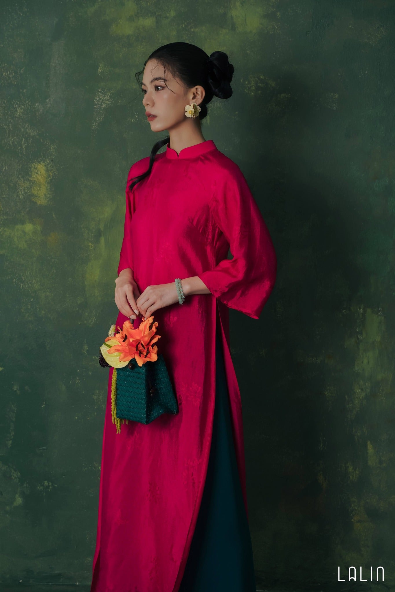 Woman in a bright pink Ao dai holding a colorful flower against a dark green background