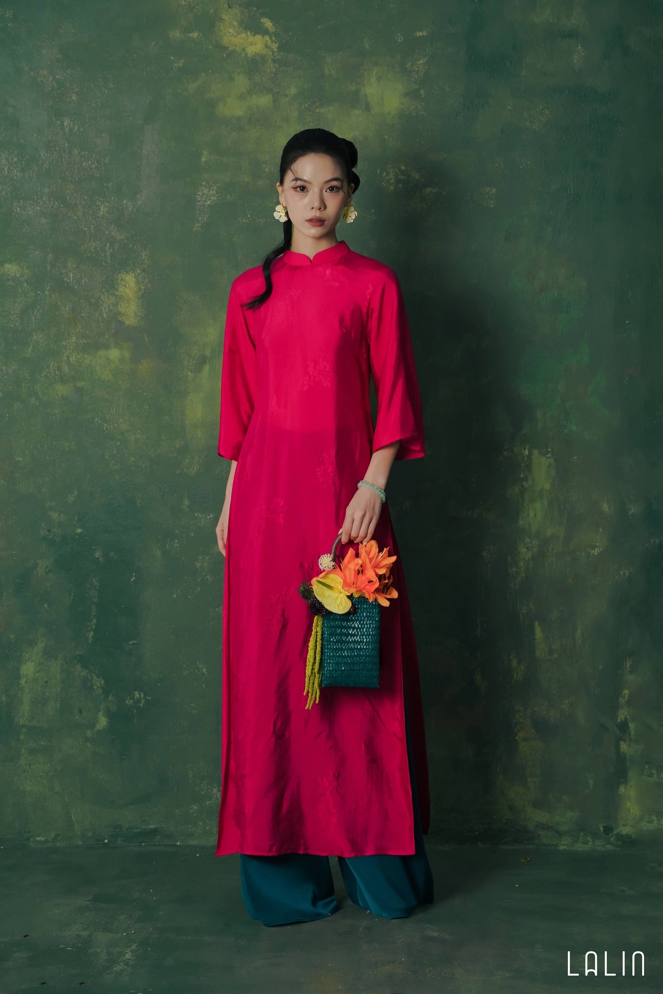 Woman in a bright pink Ao dai holding a colorful bag against a textured green wall.