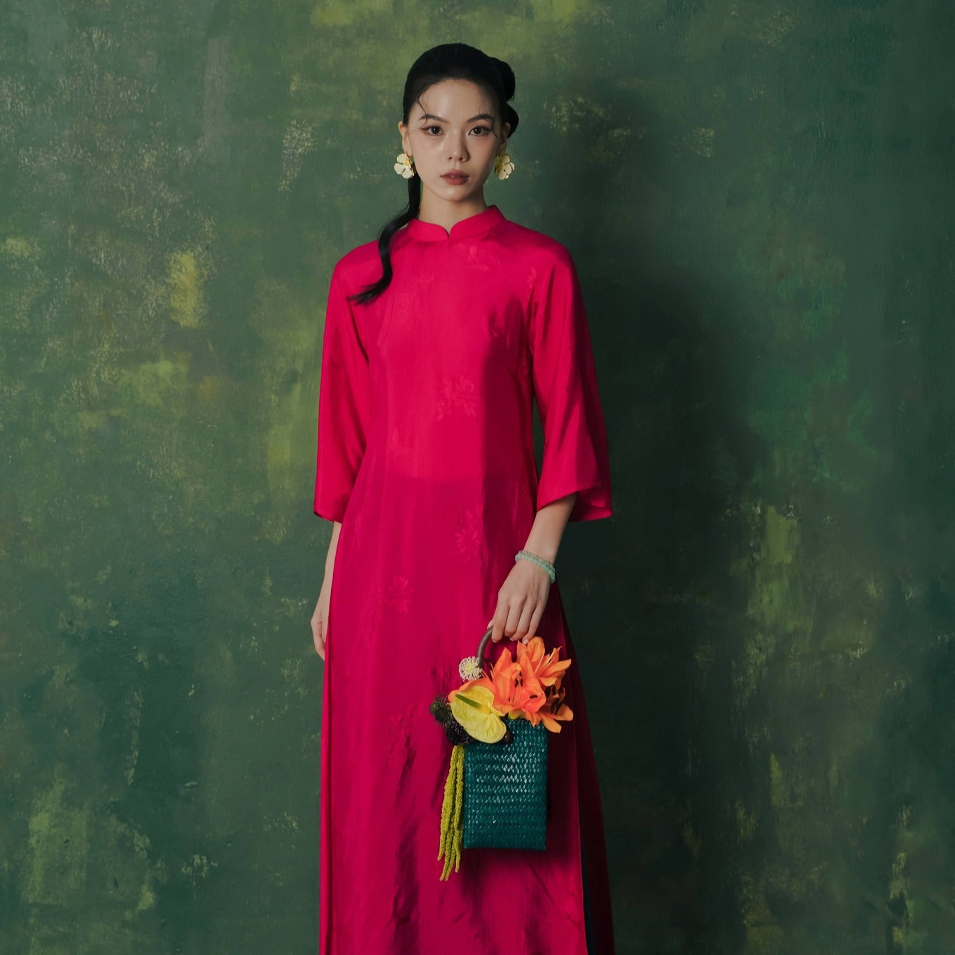 Woman in a bright pink Ao dai holding a colorful bag against a textured green background