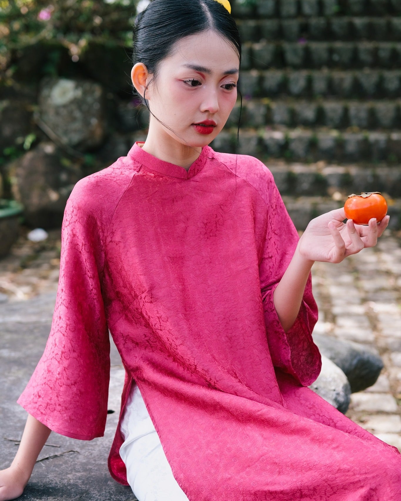 A person in a pink, patterned tunic holds a small orange fruit, sitting on stone steps outdoors.