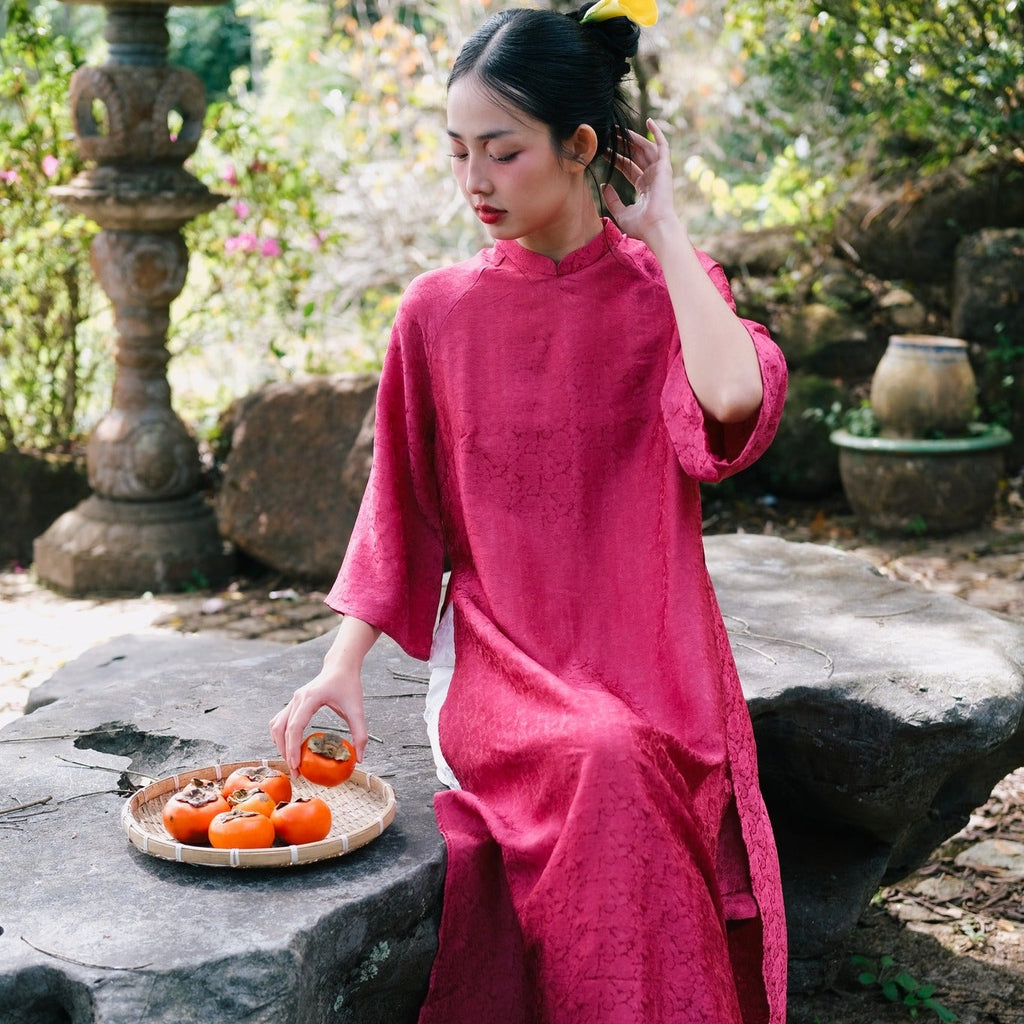 A person in a red traditional dress sits outdoors on a stone bench, reaching for a persimmon on a woven tray.