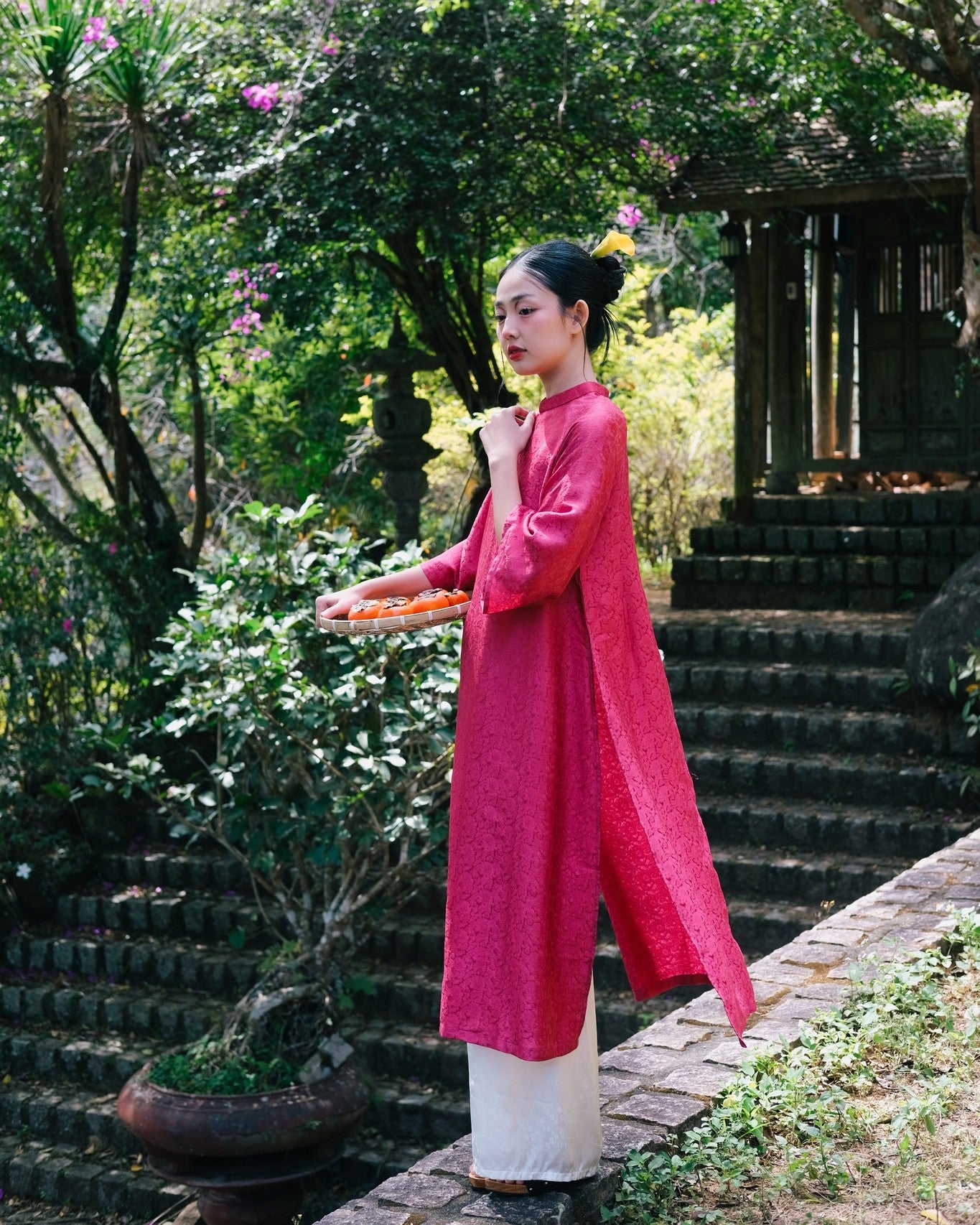 A person in a vibrant pink traditional outfit, standing outdoors in a lush garden, holding a tray with orange pastries.