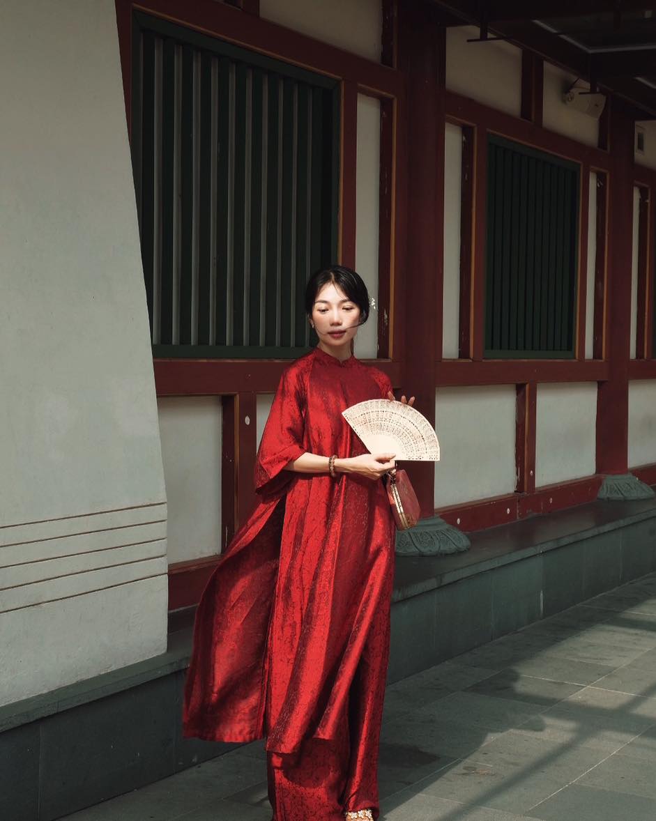A woman wearing a flowing red dress and holding a fan, standing by a traditional building with wooden elements in the background.