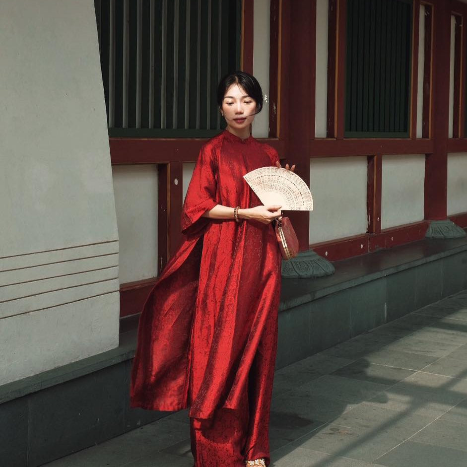 A woman wearing a flowing red dress and holding a fan, standing by a traditional building with wooden elements in the background.