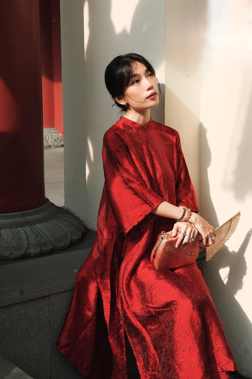 A woman in a flowing red dress sits in sunlight, holding a clutch and fan.